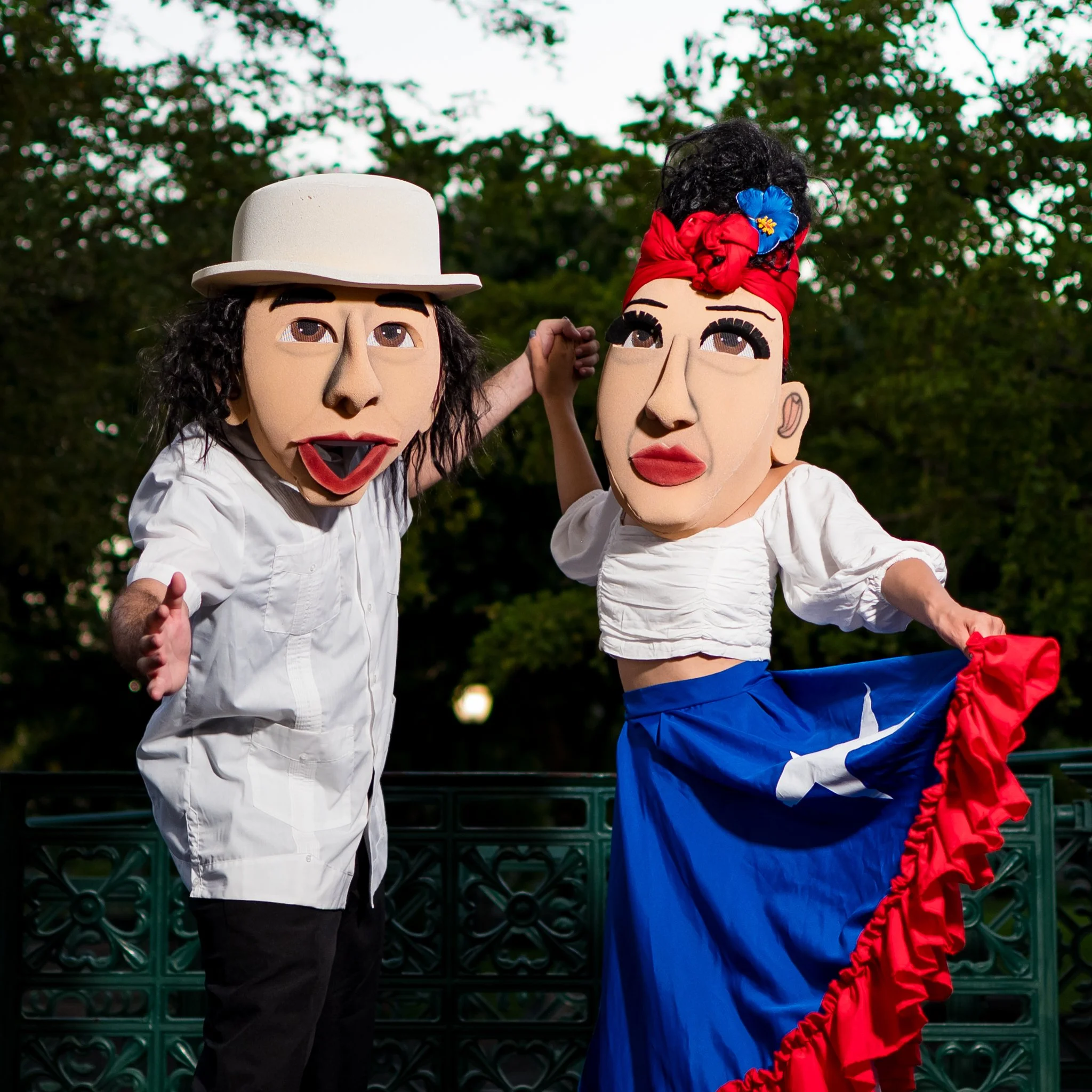 Two people wearing large cartoon-style masks, one with a white hat and long dark hair, the other with a red headscarf and blue skirt, standing outdoors in front of greenery.