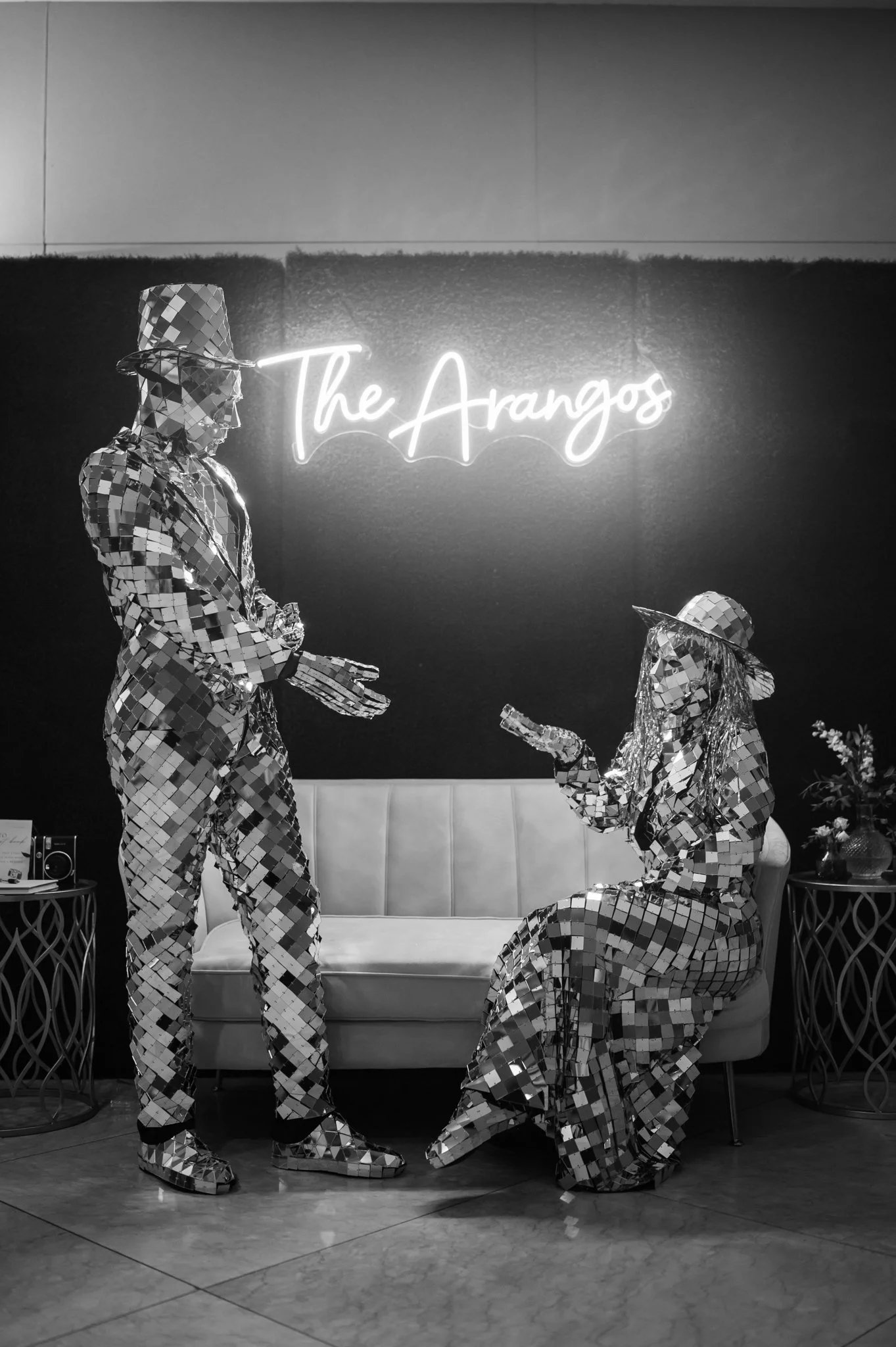 Two people dressed in mirrored costumes and hats, one standing and one sitting, in front of a neon sign that says 'The Arangos' at a stylish interior venue.
