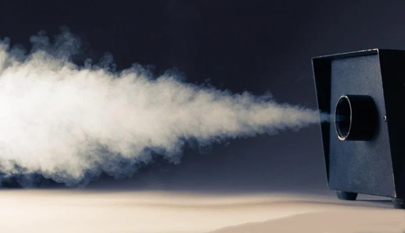 A black portable fog machine emitting a thick cloud of white fog or vapor to the left.