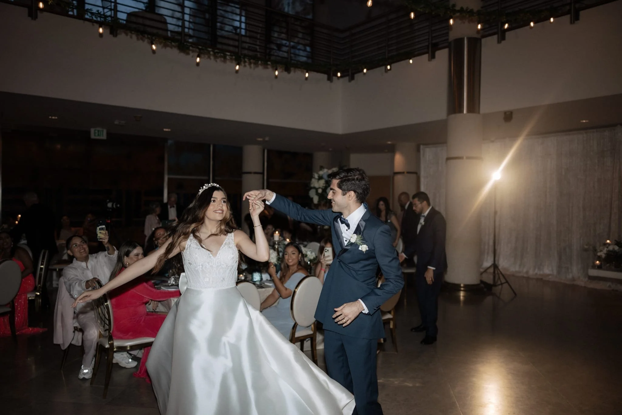 A bride and groom dancing at their wedding reception, surrounded by seated guests in a decorated indoor venue with string lights overhead.