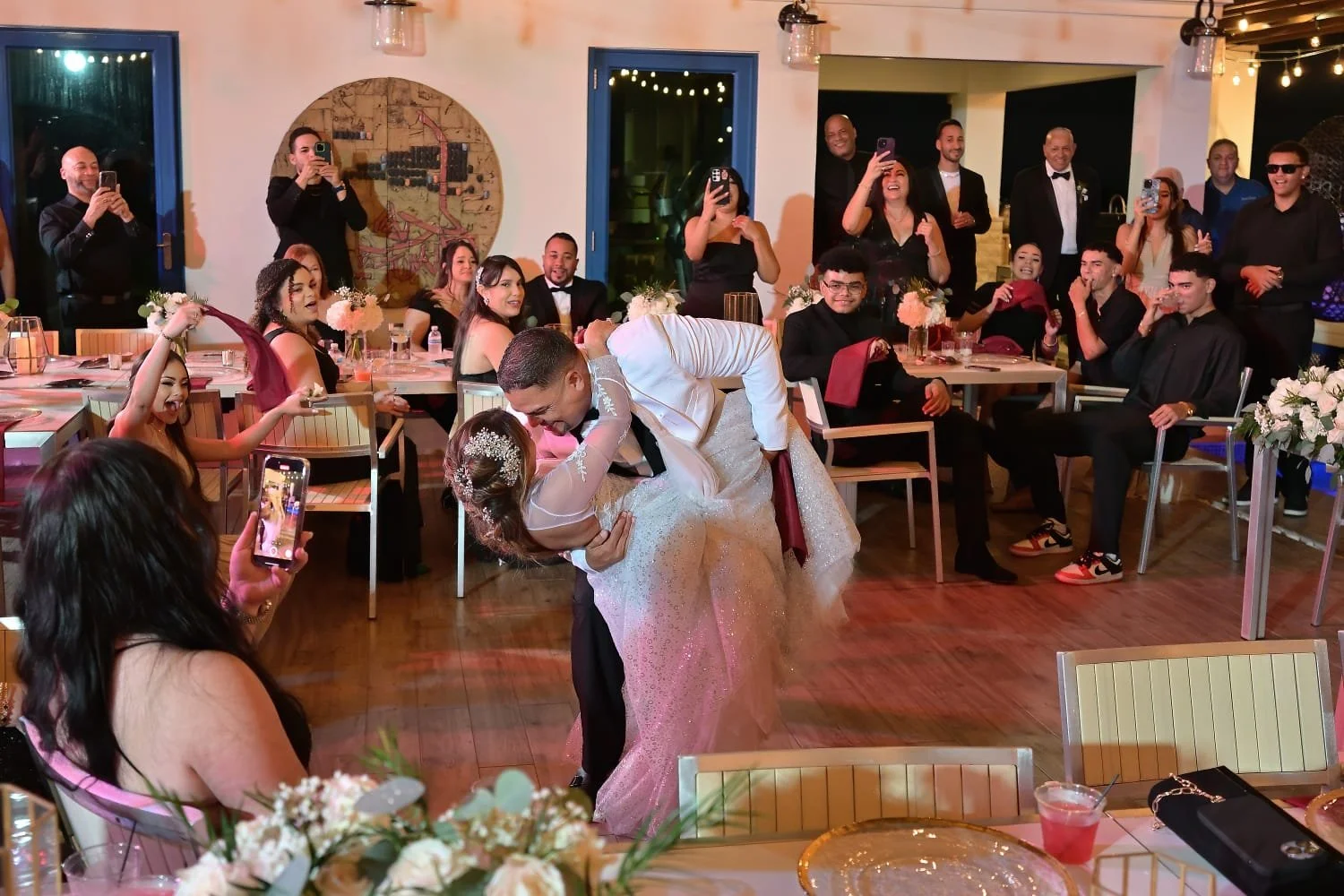 A newlywed couple shares a dance at their wedding reception, with guests watching and taking photos around them. The groom is dipping the bride, who is wearing a white wedding gown, while the guests are seated at tables and standing nearby, smiling and capturing the moment.