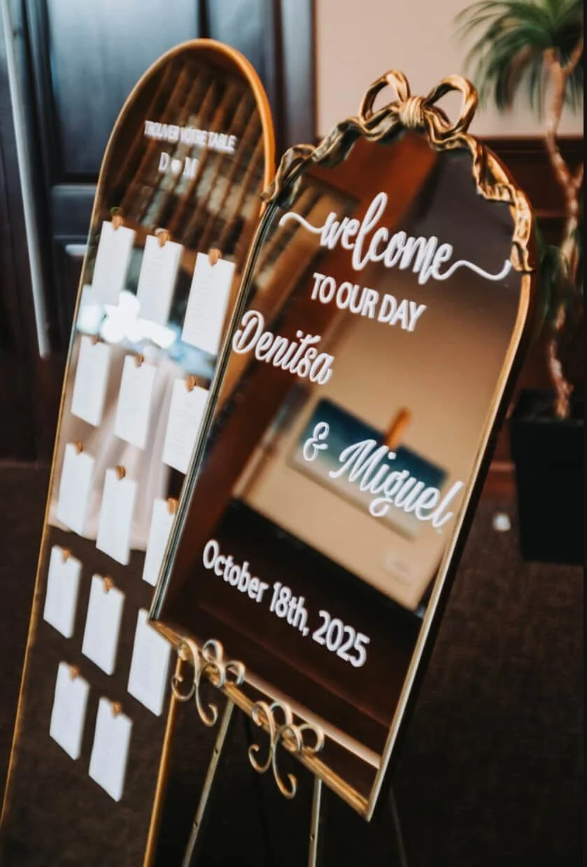 Wedding seating chart and welcome sign on gold display stands with a decorative ribbon at the top, with a plant visible in the background.