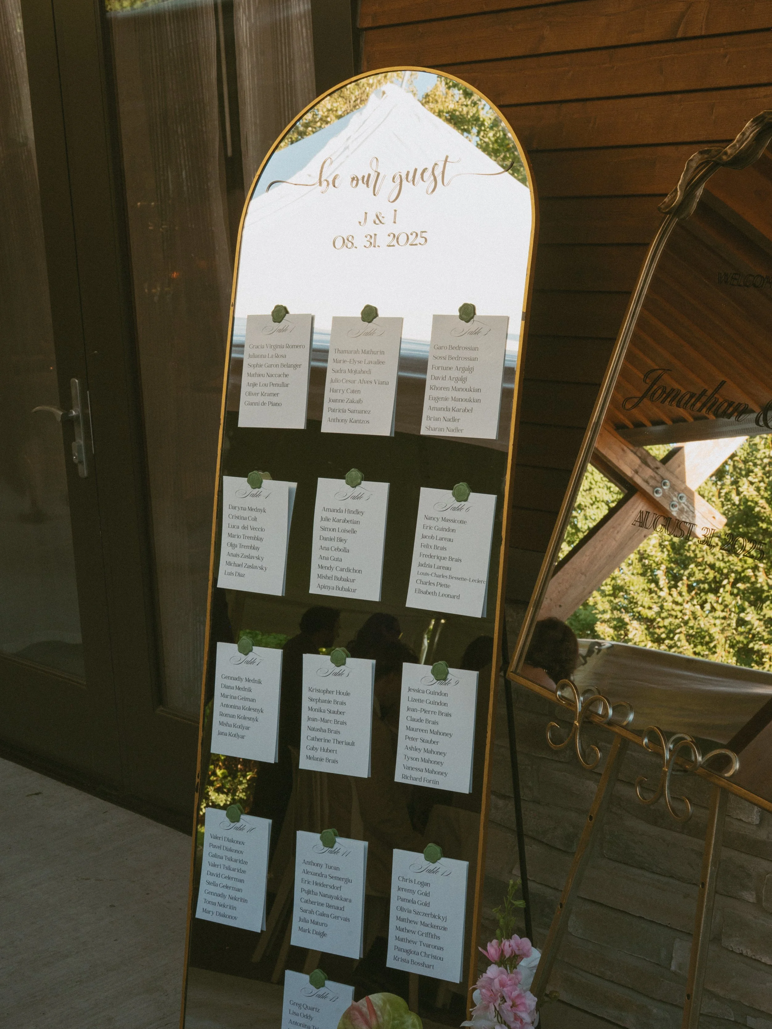 Wedding seating chart on a gold-framed mirror with multiple tabs, each listing names of guests for different tables, placed outdoors with trees and a wooden structure in the background.