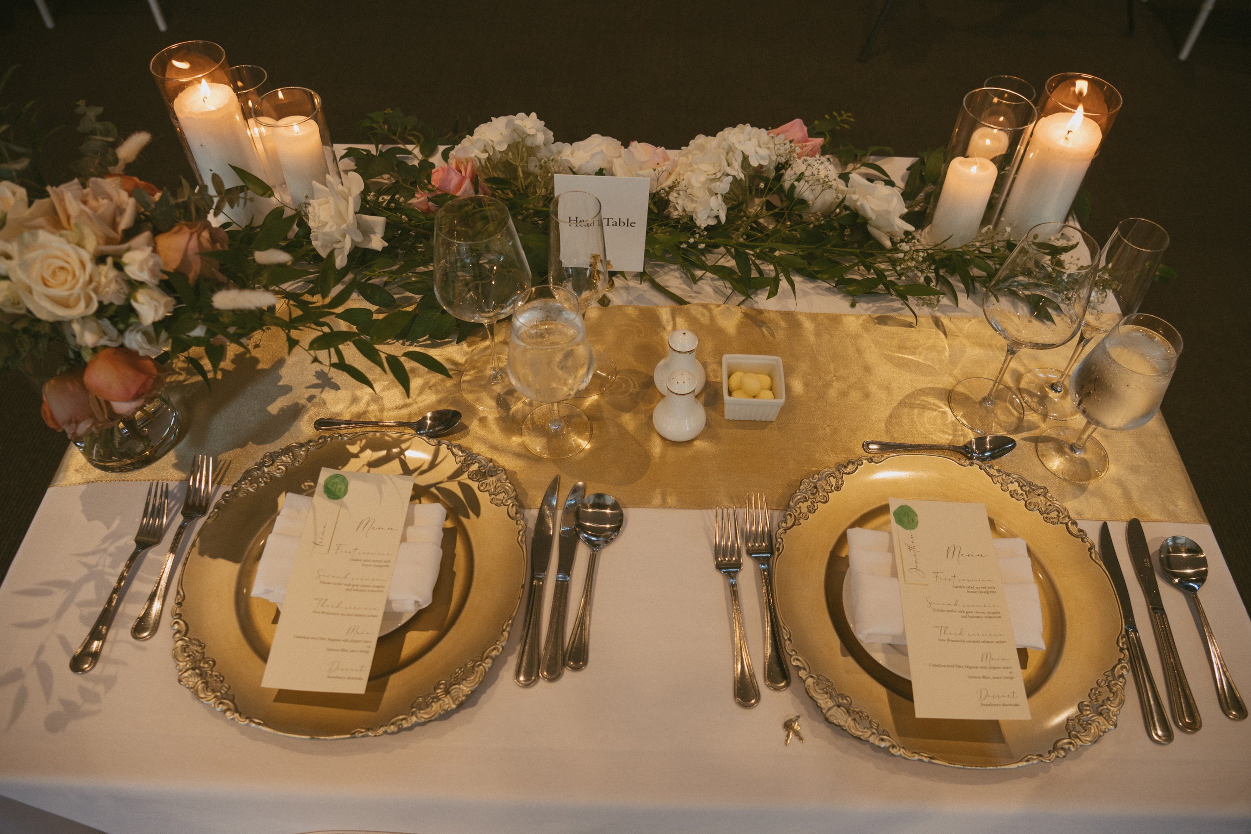 Elegant wedding or formal event table setting with gold chargers, white napkins, printed menus, multiple wine glasses, silverware, a floral centerpiece with white and blush roses and greenery, and lit candles.