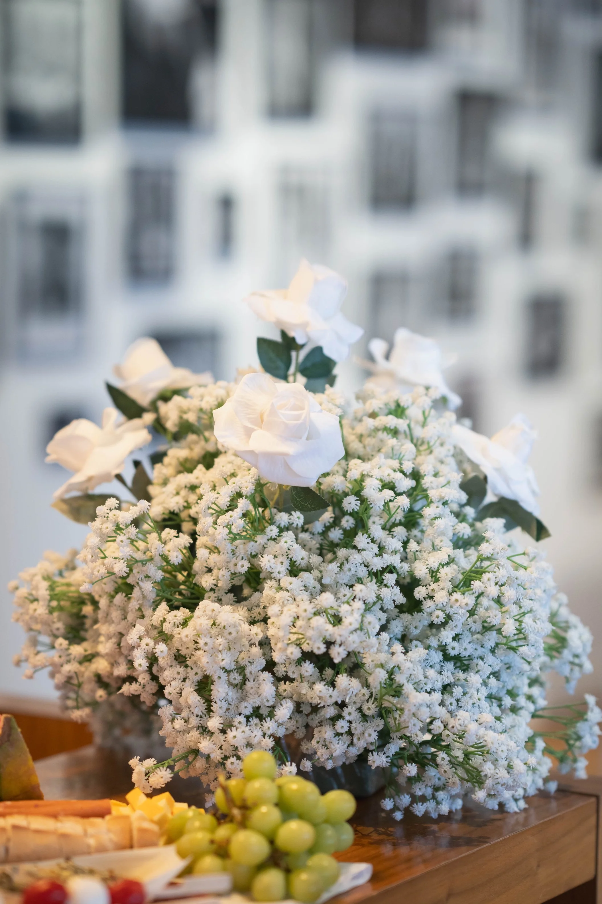 A floral bouquet of white roses and white daisy-like flowers on a wooden table, with green grapes and cheese in the foreground, and blurred window backgrounds.