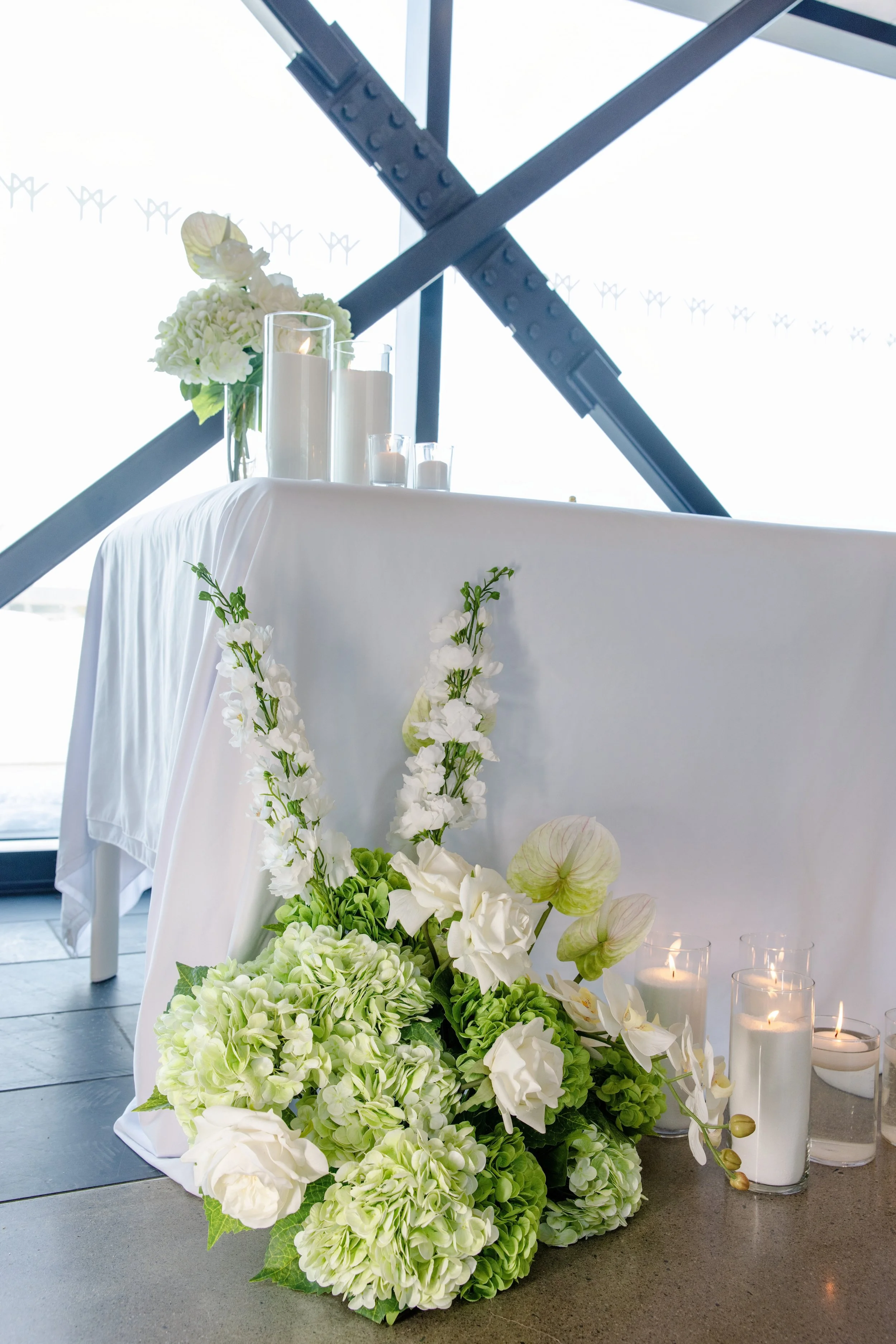 White floral arrangement with candles on a white-covered table in front of large windows and structural beams.