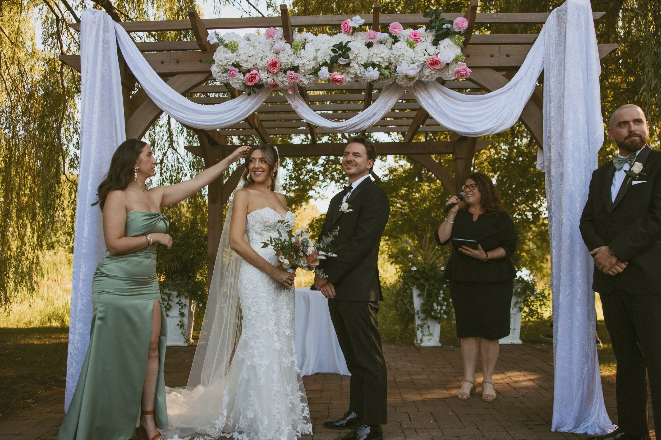 A wedding ceremony taking place outdoors under a decorated wooden arch with pink and white flowers and white drapes. The bride, in a white lace wedding gown, holds a bouquet of flowers. The groom, in a black tuxedo, stands across from her. A woman in