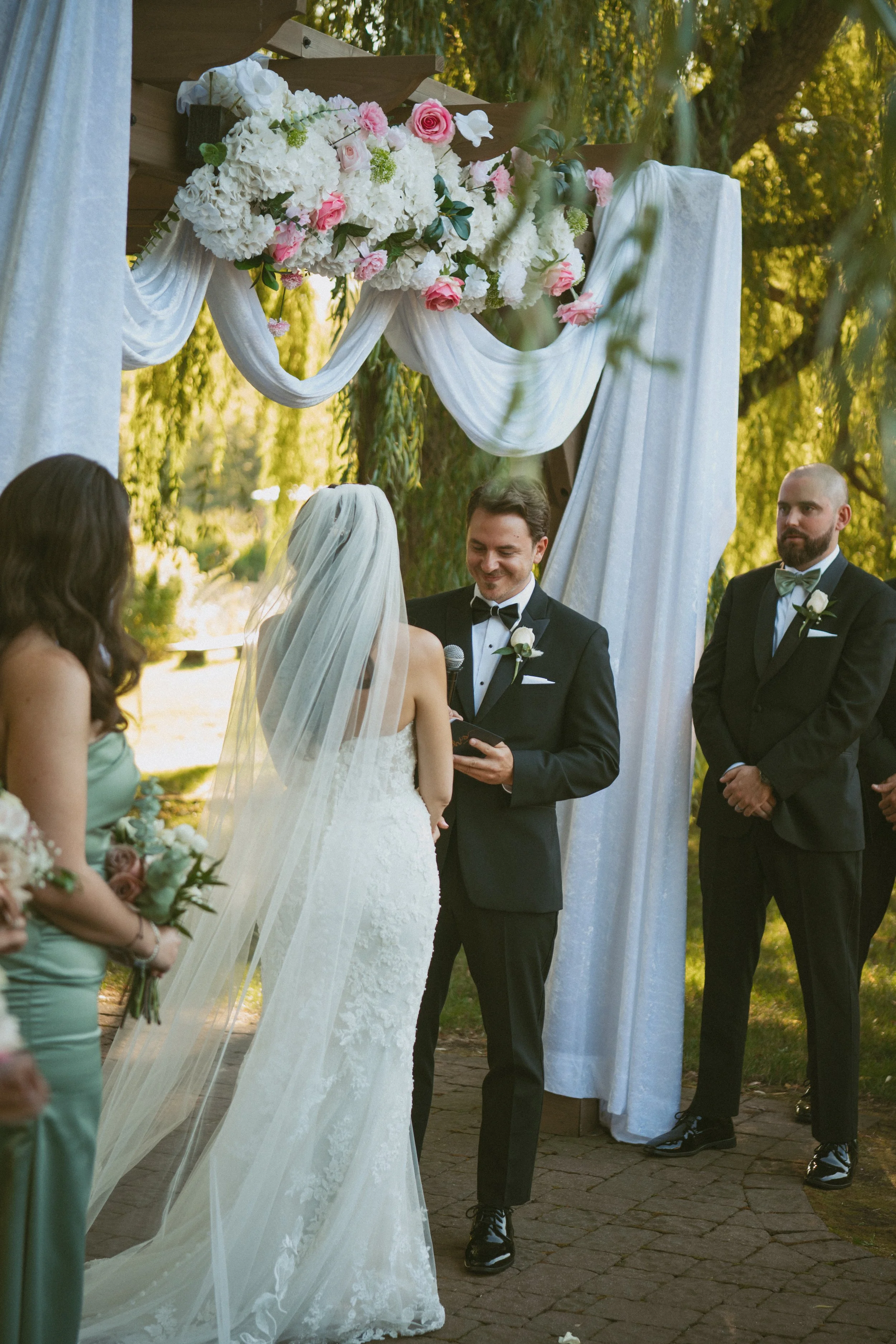 A wedding ceremony outdoors with a bride in a white lace gown and veil, standing in front of a groom in a black tuxedo, exchanging vows. Two bridesmaids and a groomsman are present, with the groomsman standing to the right. A floral arch with pink an