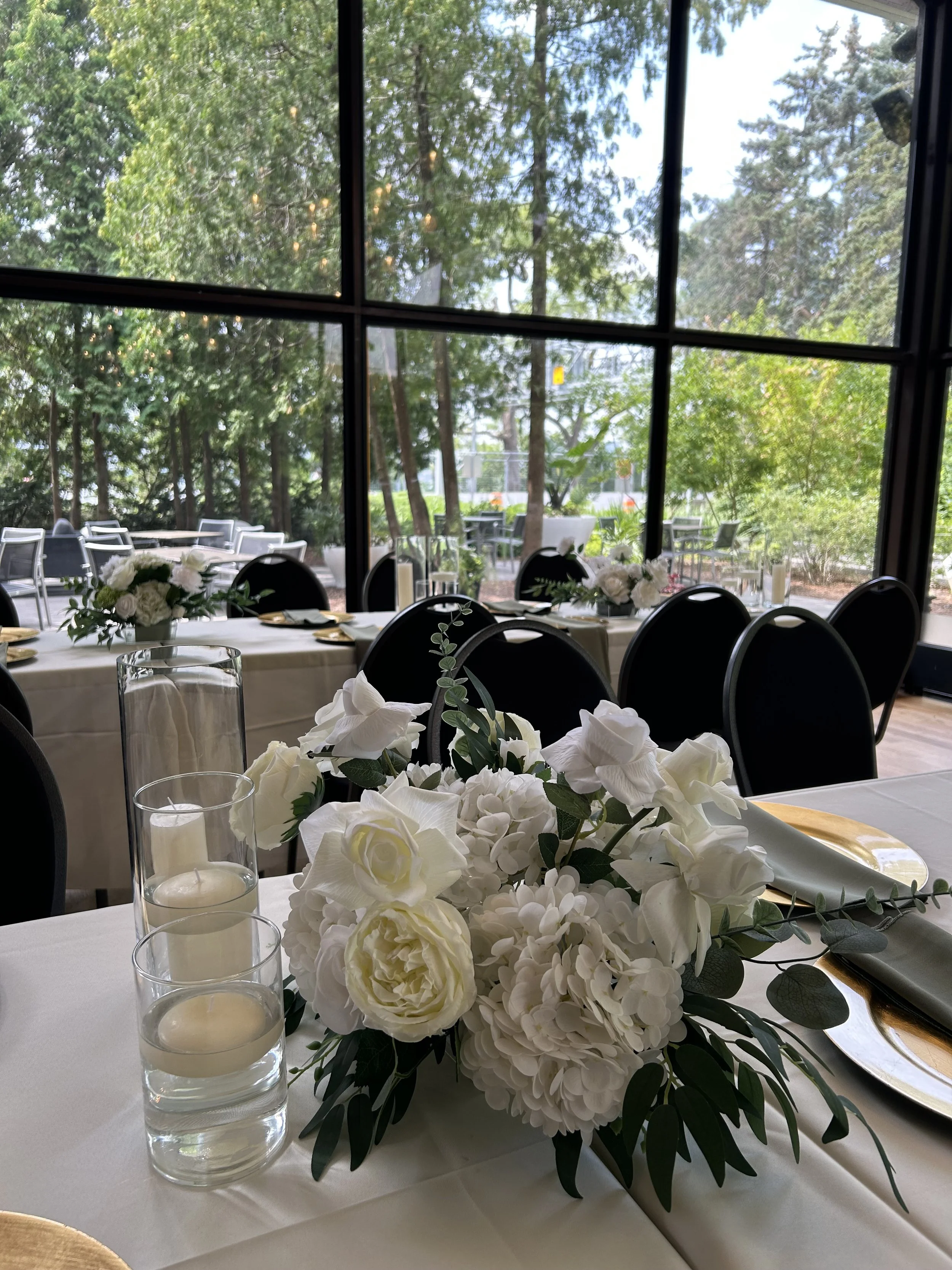 Elegant table setup with white floral centerpiece, candles, and gold-rimmed plates in a bright room with large windows overlooking a garden.