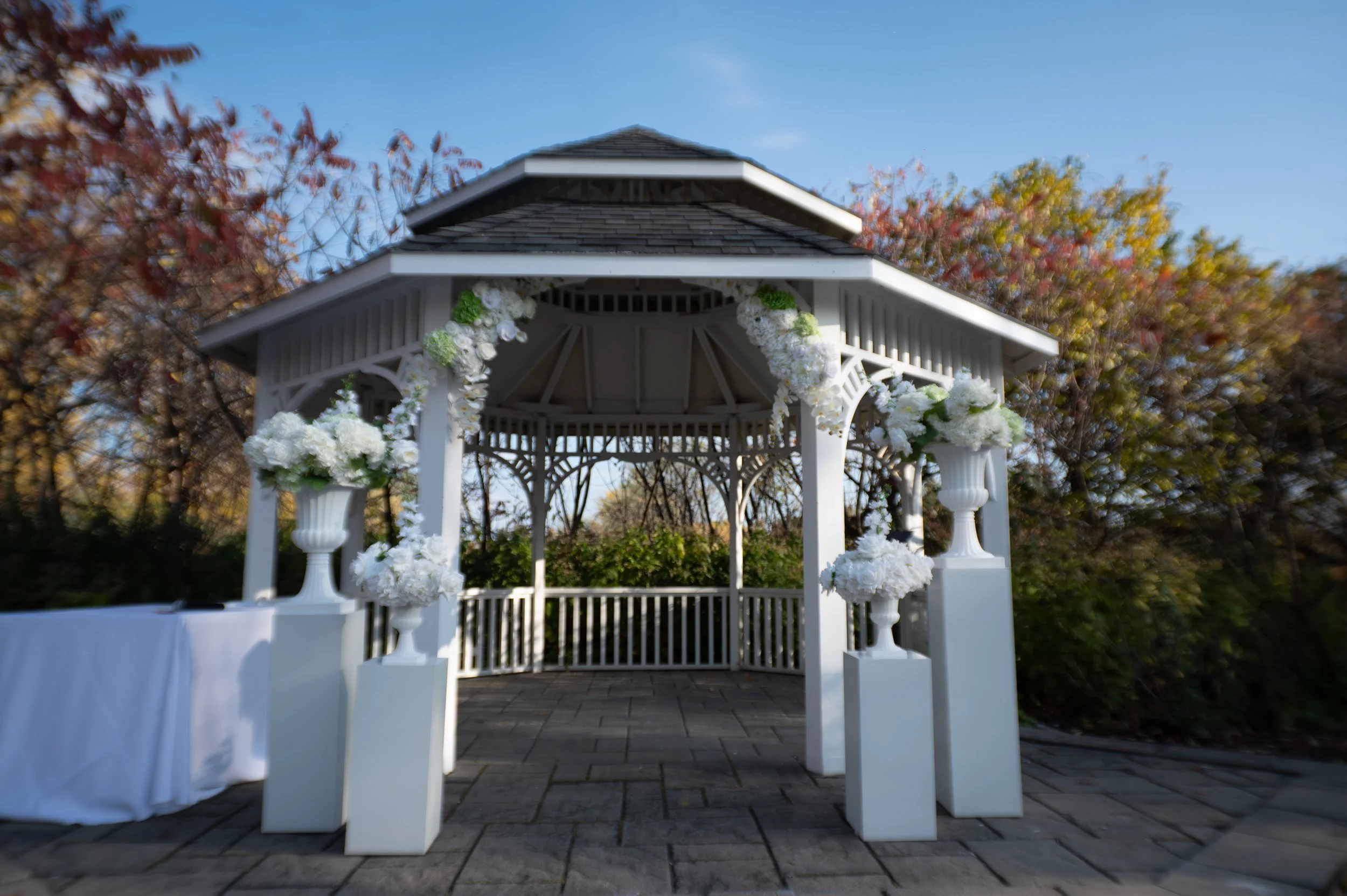 A white decorative wedding gazebo decorated with white flowers and greenery, set outdoors on a paved area with trees and colorful autumn foliage in the background.