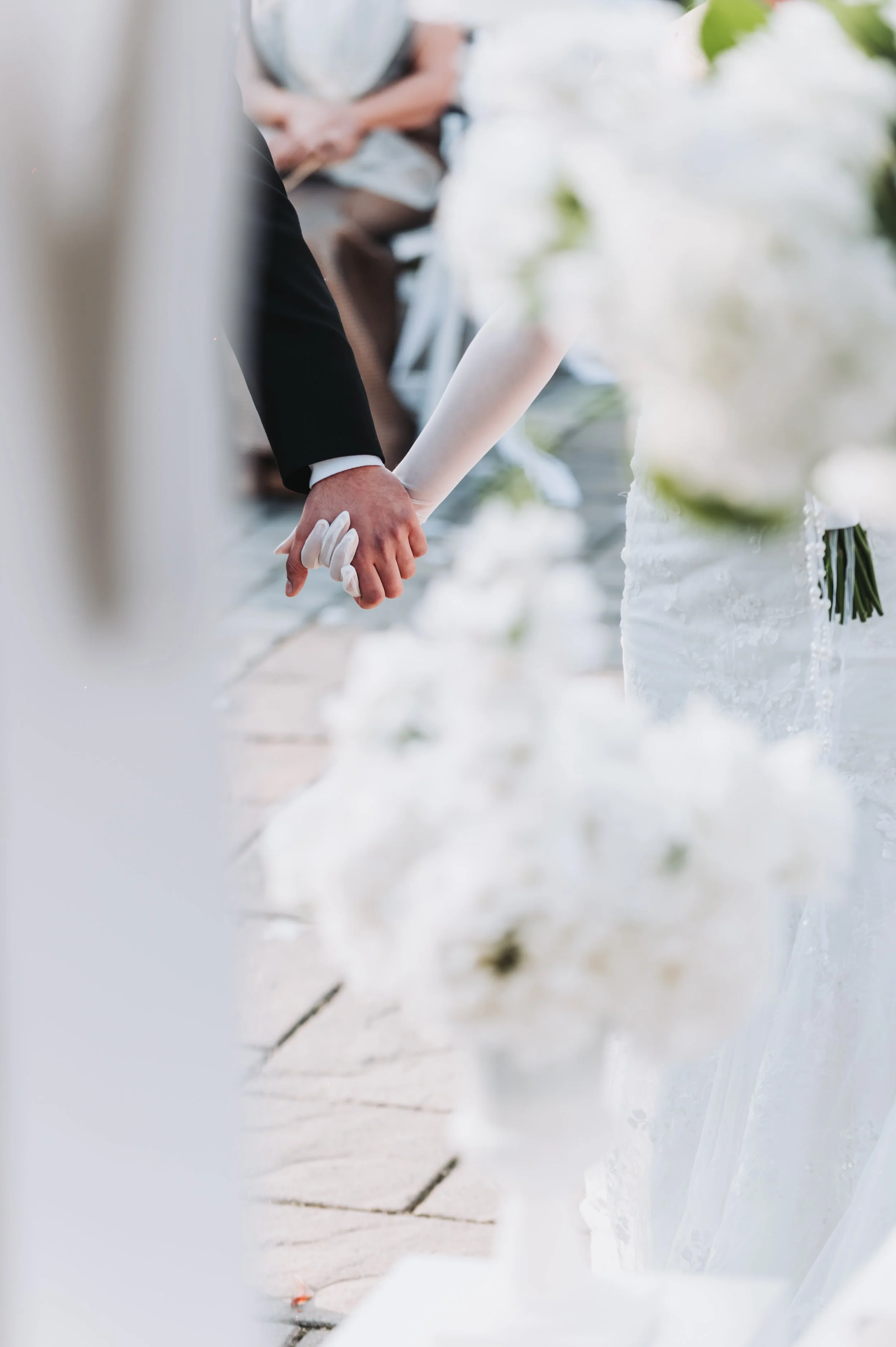 A bride and groom holding hands at their wedding ceremony, with the bride wearing a lace wedding dress and holding a bouquet of white flowers.