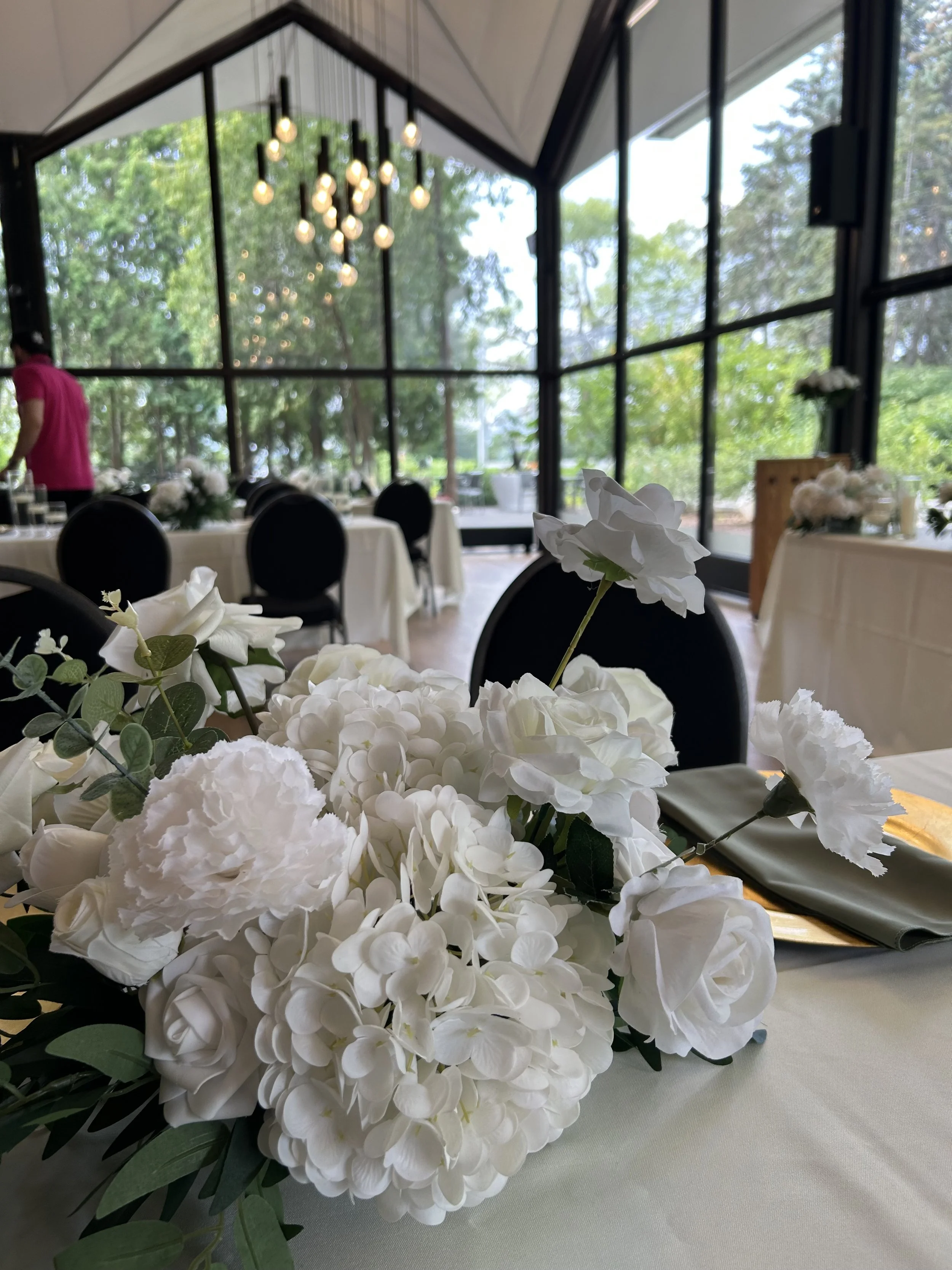 A close-up of a white floral centerpiece on a table at a decorated event venue with large glass windows and modern hanging lights.