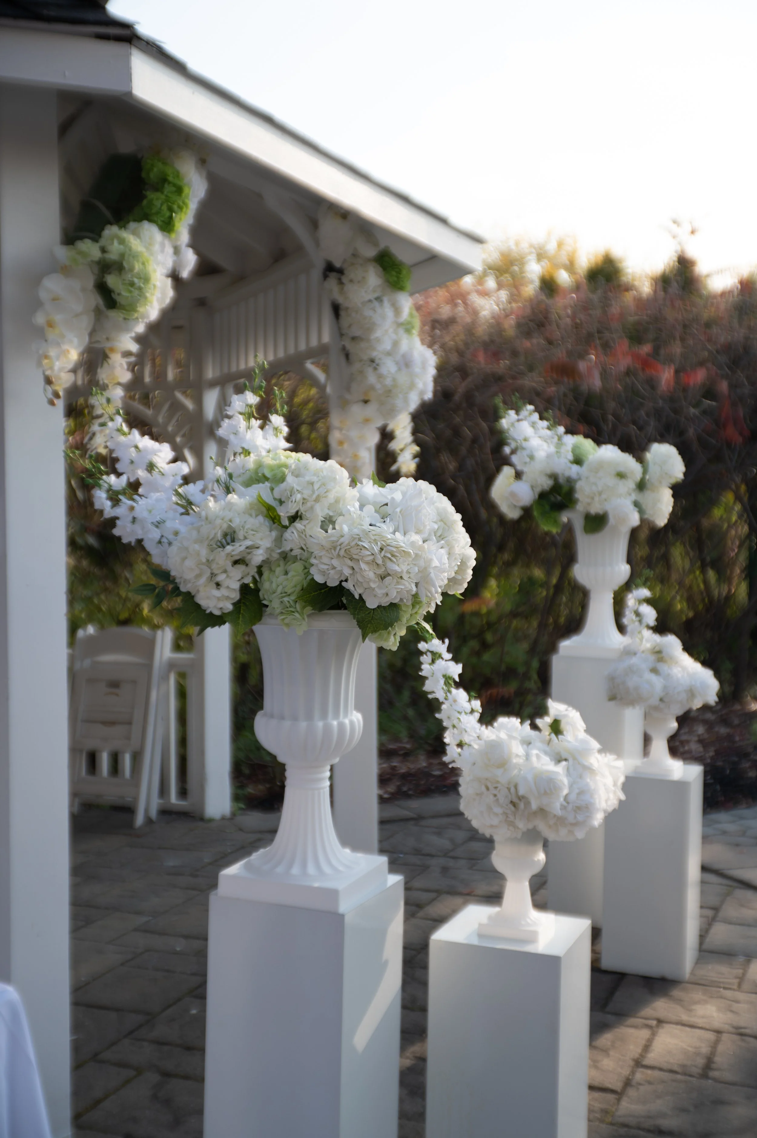 White floral arrangements in tall vases on white pedestals at an outdoor event with a gazebo and a garden background.