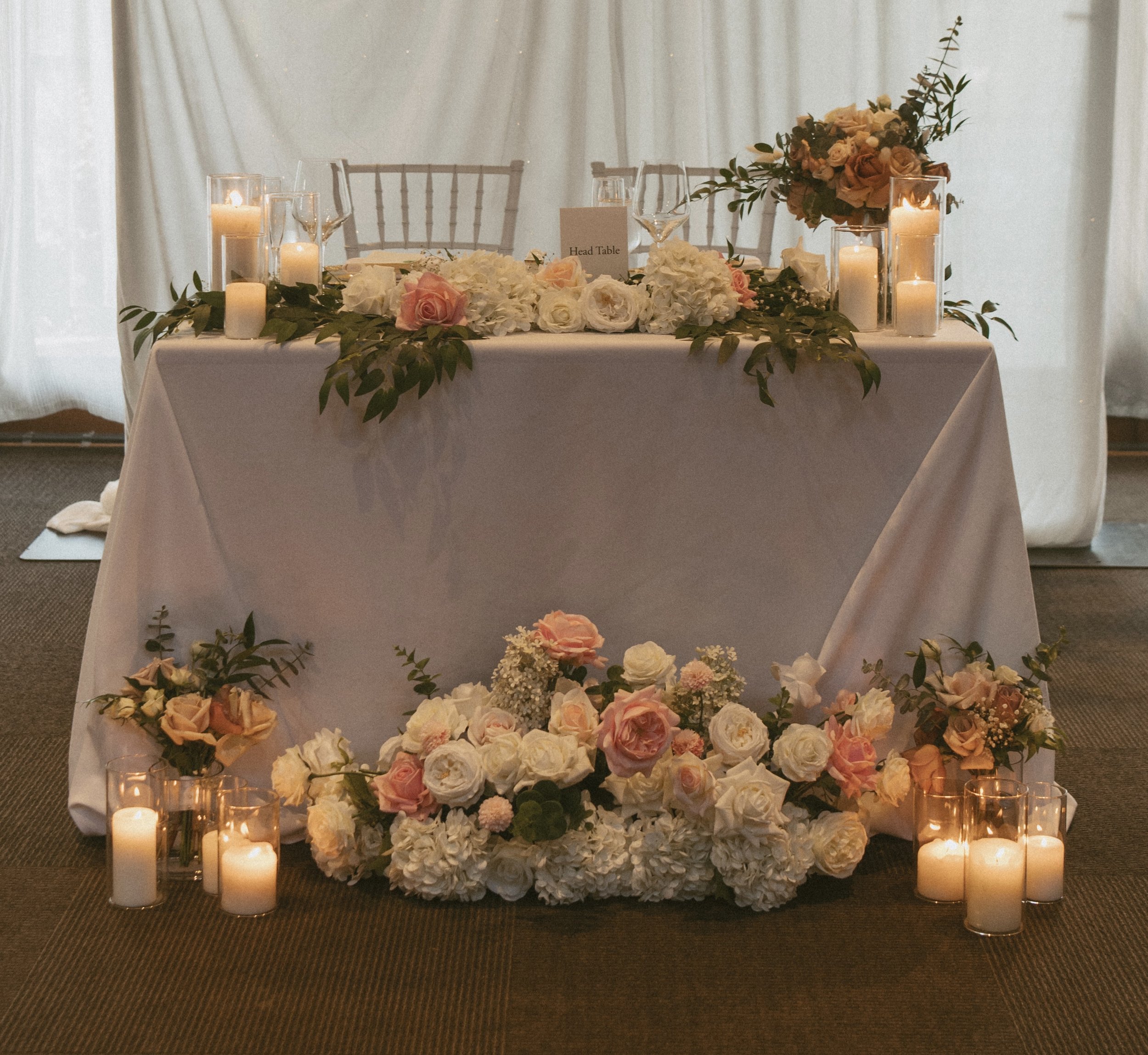 A decorated wedding or event table with floral arrangements, candles, and a sign that reads 'Head Table'. Flowers are mainly white and pink, with greenery accents. The table is covered with a white tablecloth. The background features white curtains.