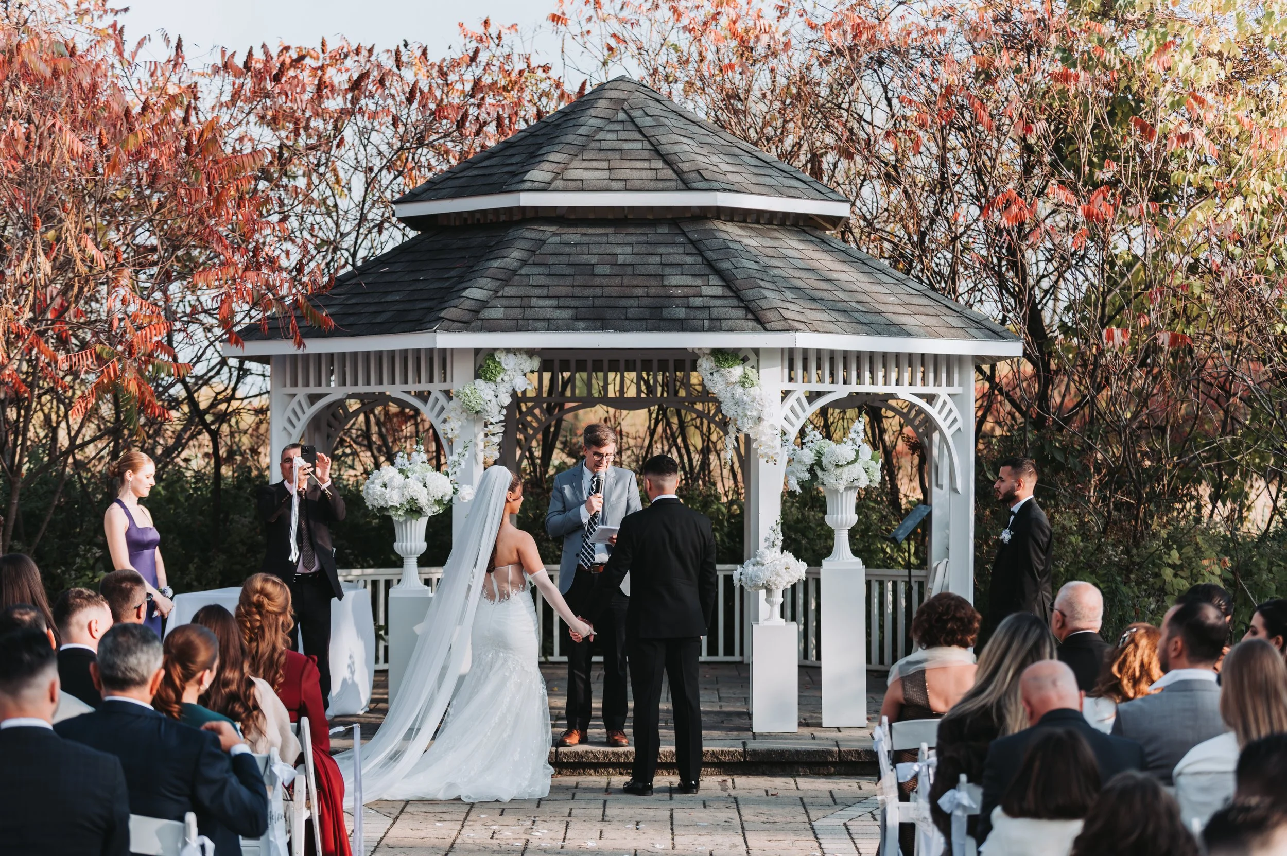 A wedding ceremony taking place outdoors under a gazebo decorated with white flowers, with the bride and groom holding hands and standing before the officiant, while guests watch.