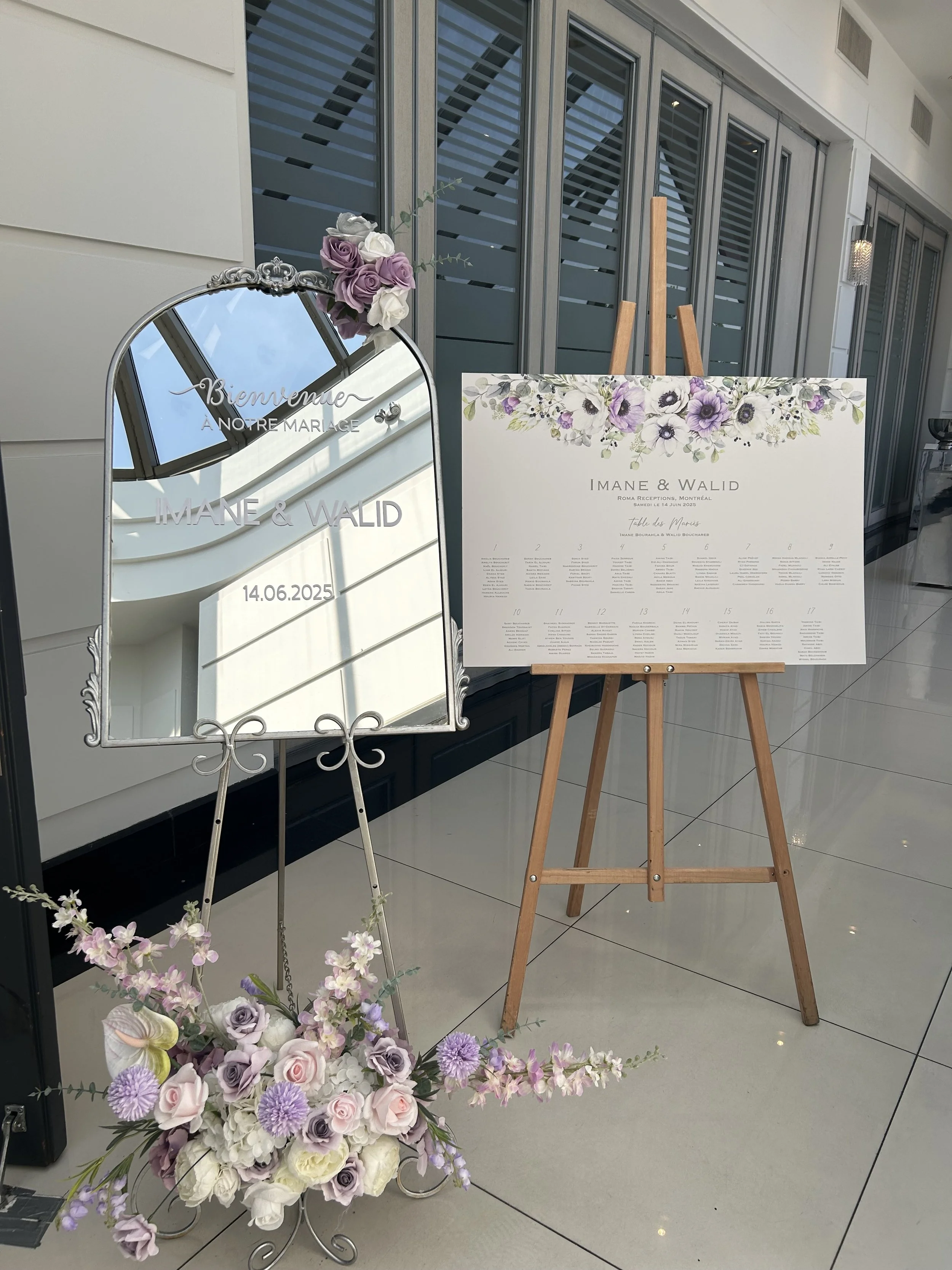 Wedding reception signage with a decorative floral border and a wedding seating chart on a wooden easel, accompanied by a mirror framed with flowers, set in a modern indoor space.
