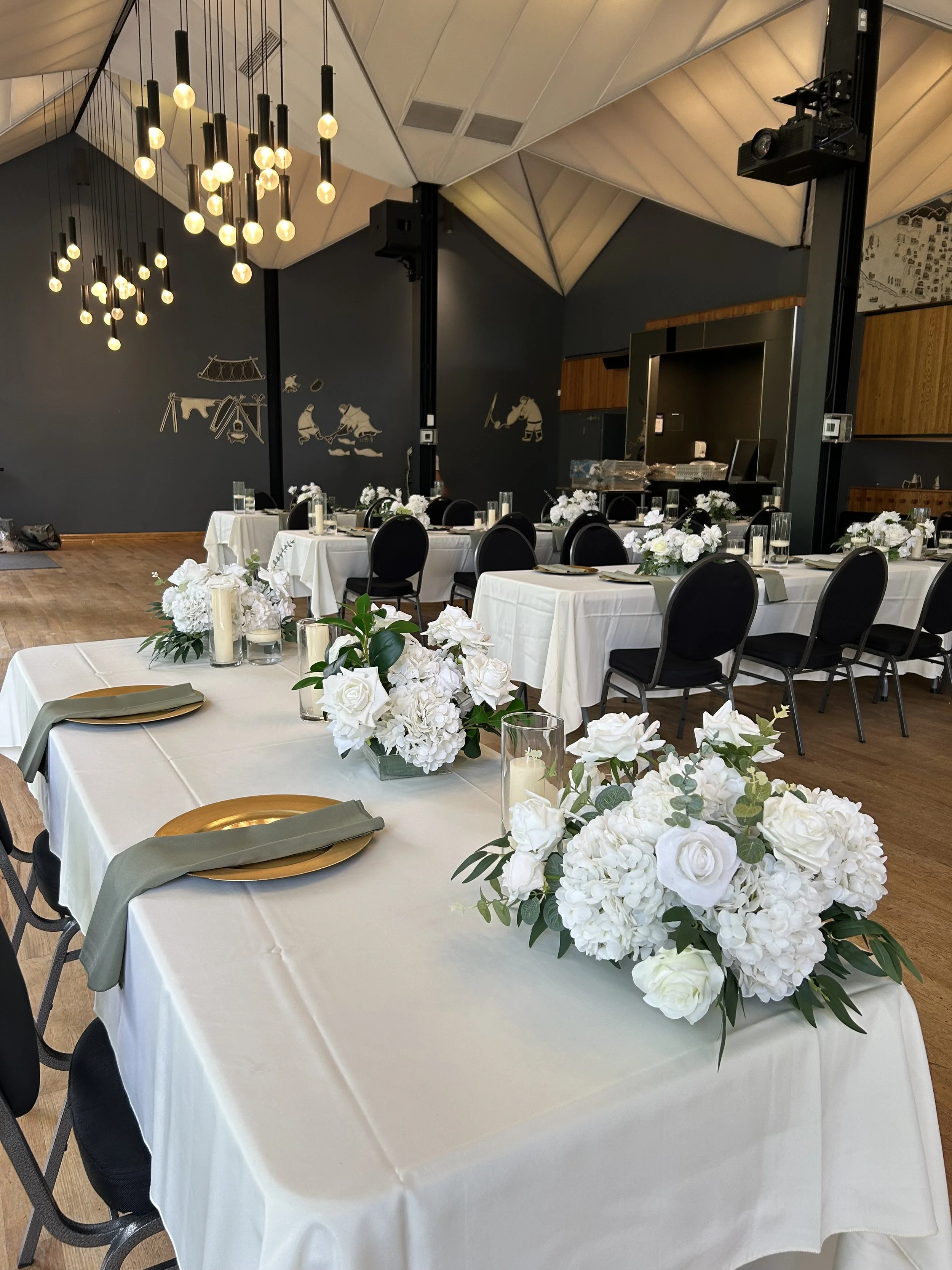 Elegant banquet hall with white tablecloths, floral centerpieces, black chairs, and modern hanging light fixtures.