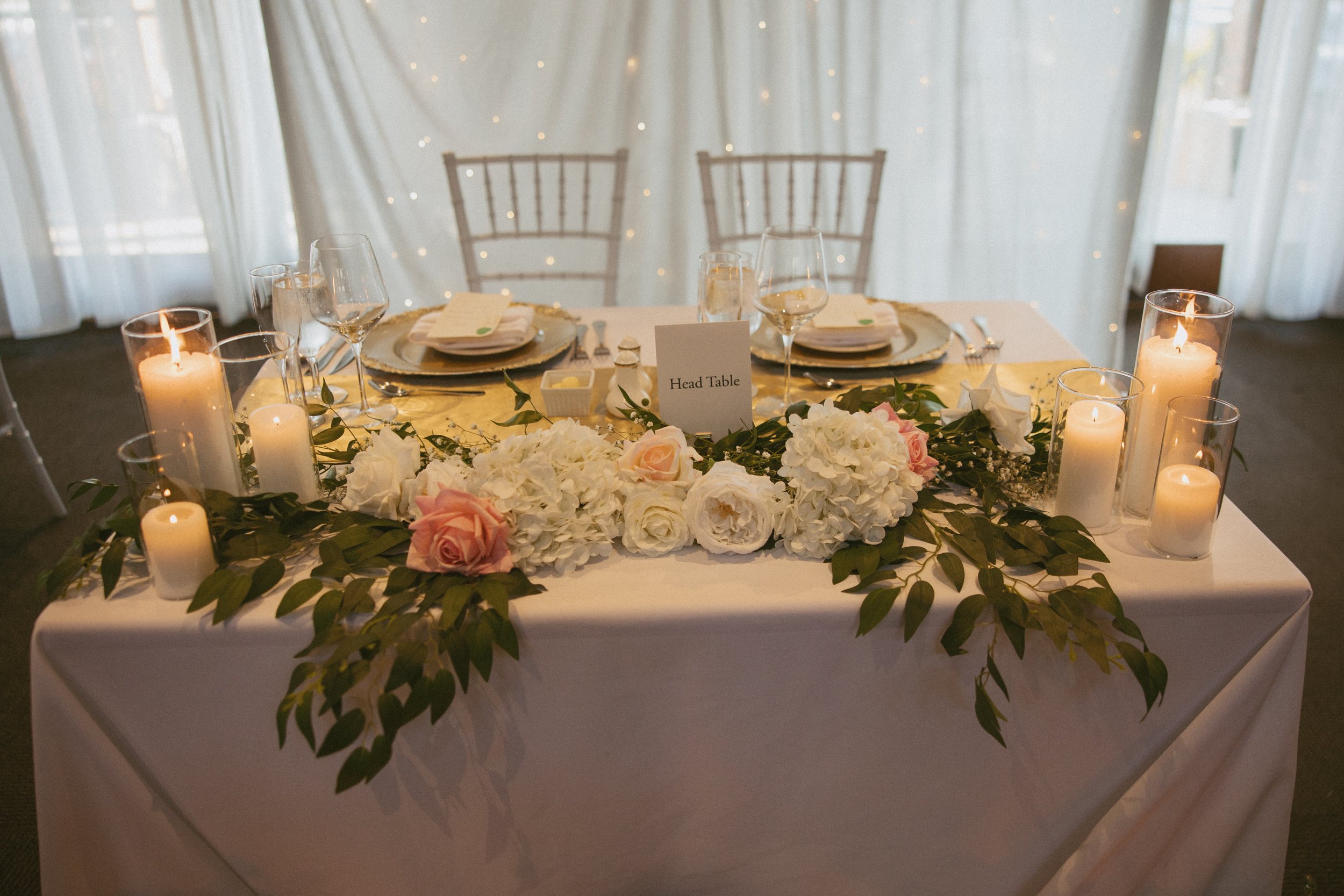 A decorated wedding reception table with white and pink floral arrangements, candles, and place settings, with a sign reading 'Head Table' in the center.
