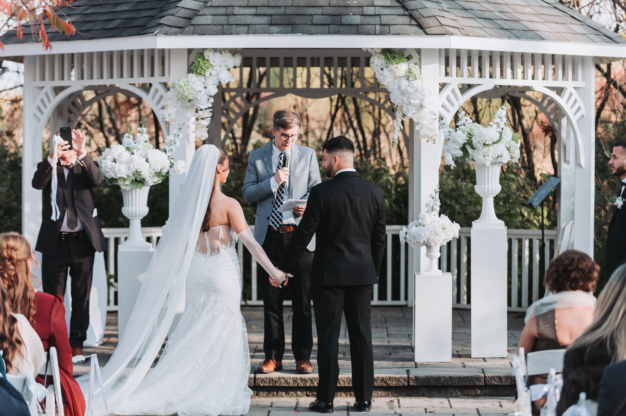 A wedding ceremony taking place outdoors under a decorated white gazebo, with a bride and groom holding hands and facing an officiant, during daytime. Guests are seated and watching, and a man is taking photos.