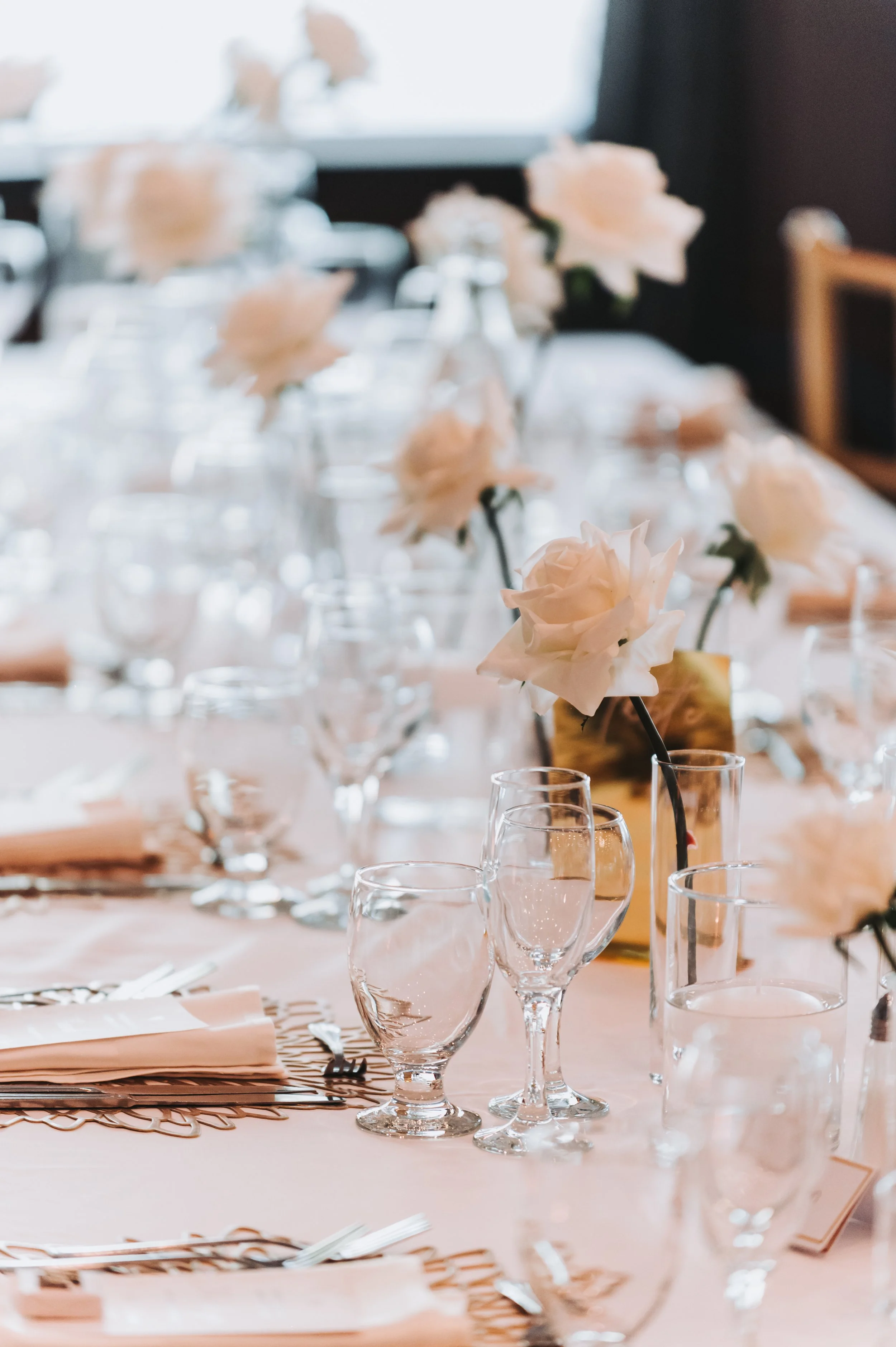 A elegantly set dining table with white roses in bottles, clear glassware, and napkins arranged on a white tablecloth for a formal event.