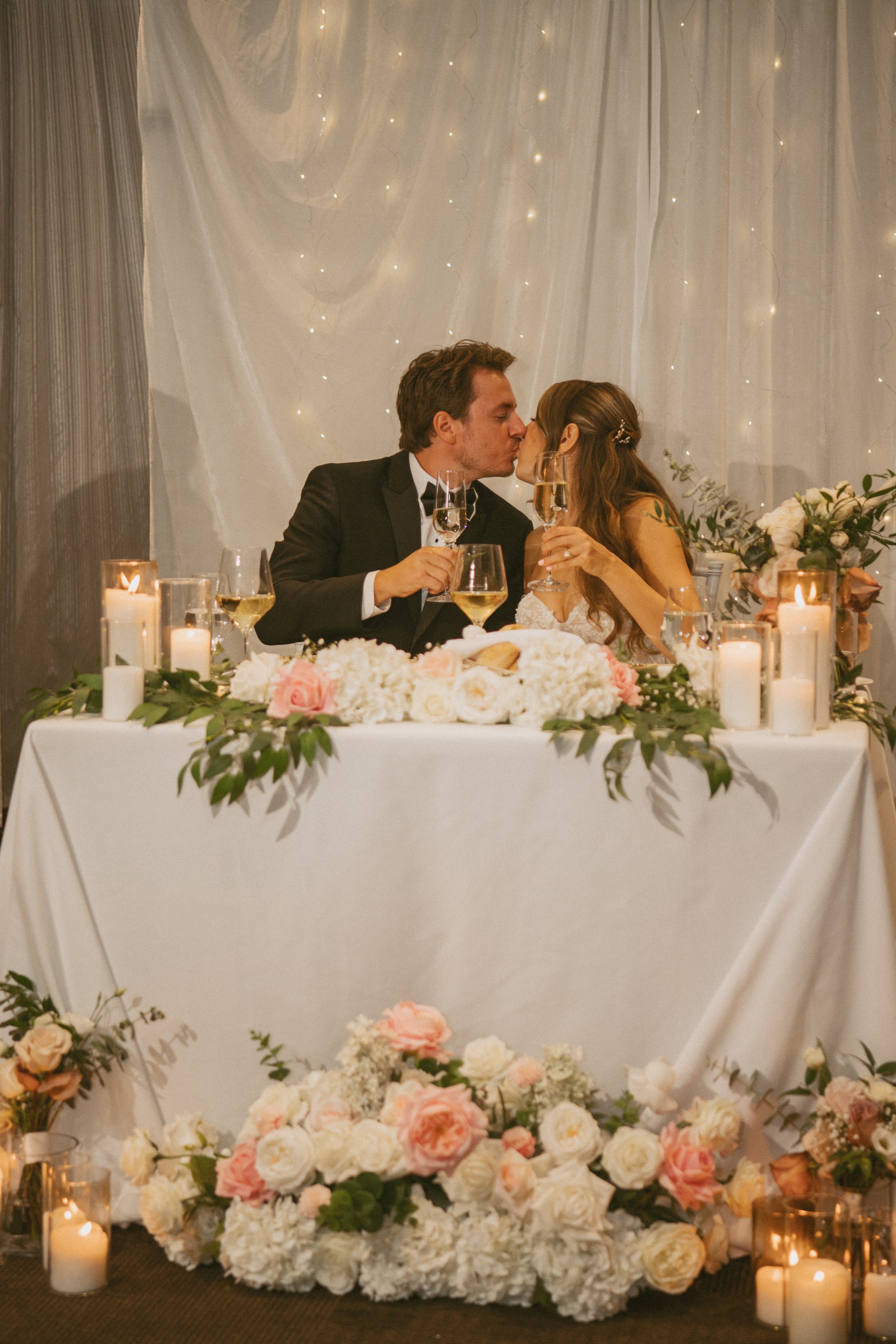 A bride and groom kiss at their wedding reception table decorated with white and pink flowers and lit candles.