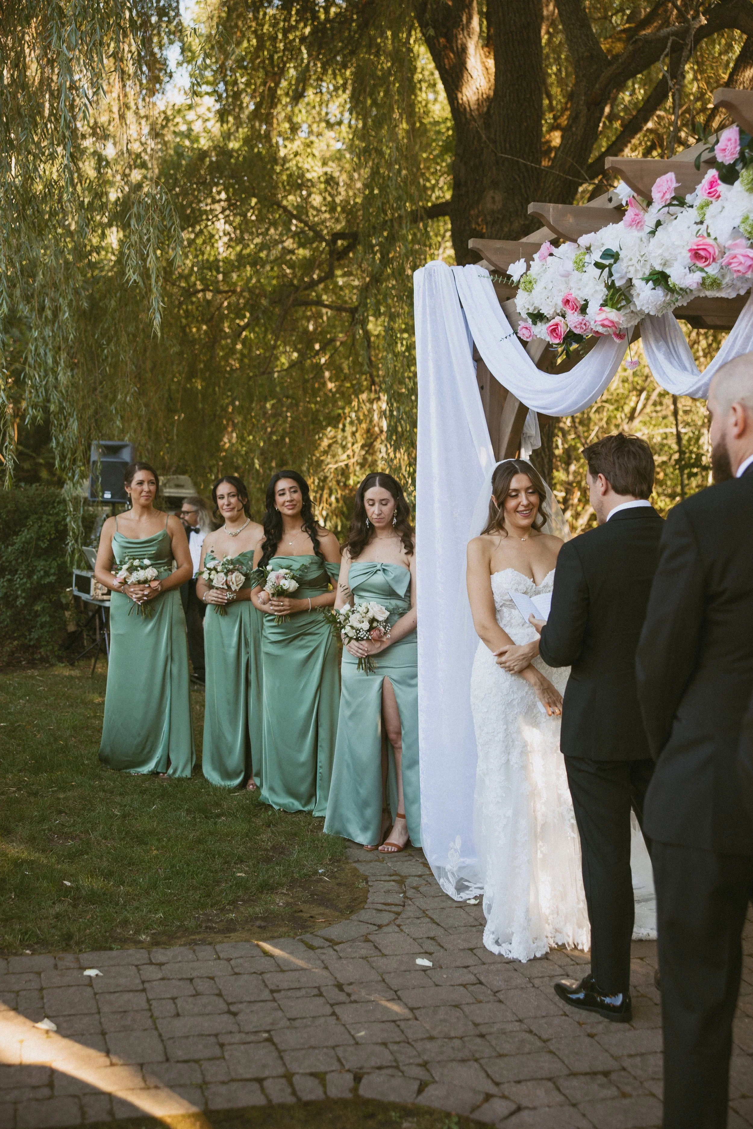 Wedding ceremony outdoors with bride and groom exchanging vows, officiant, and bridesmaids in green dresses standing in line under wooden arch decorated with white drapery and pink flowers.
