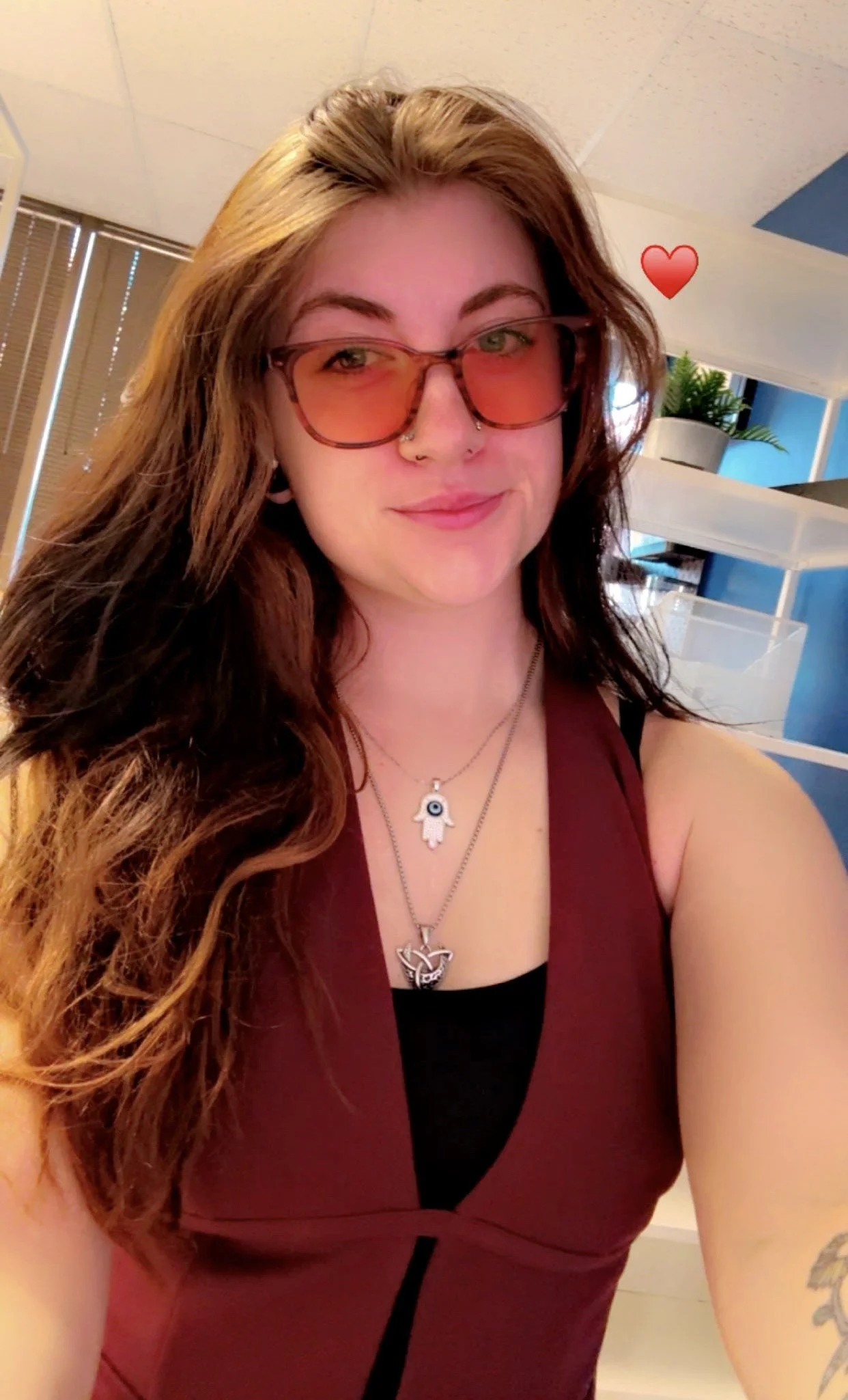 A woman with long, wavy brown hair wearing pink sunglasses, a burgundy sleeveless top, and two necklaces, one with a Hamsa hand and another with a heart-shaped pendant, taking a selfie indoors with a red heart sticker added in the image.