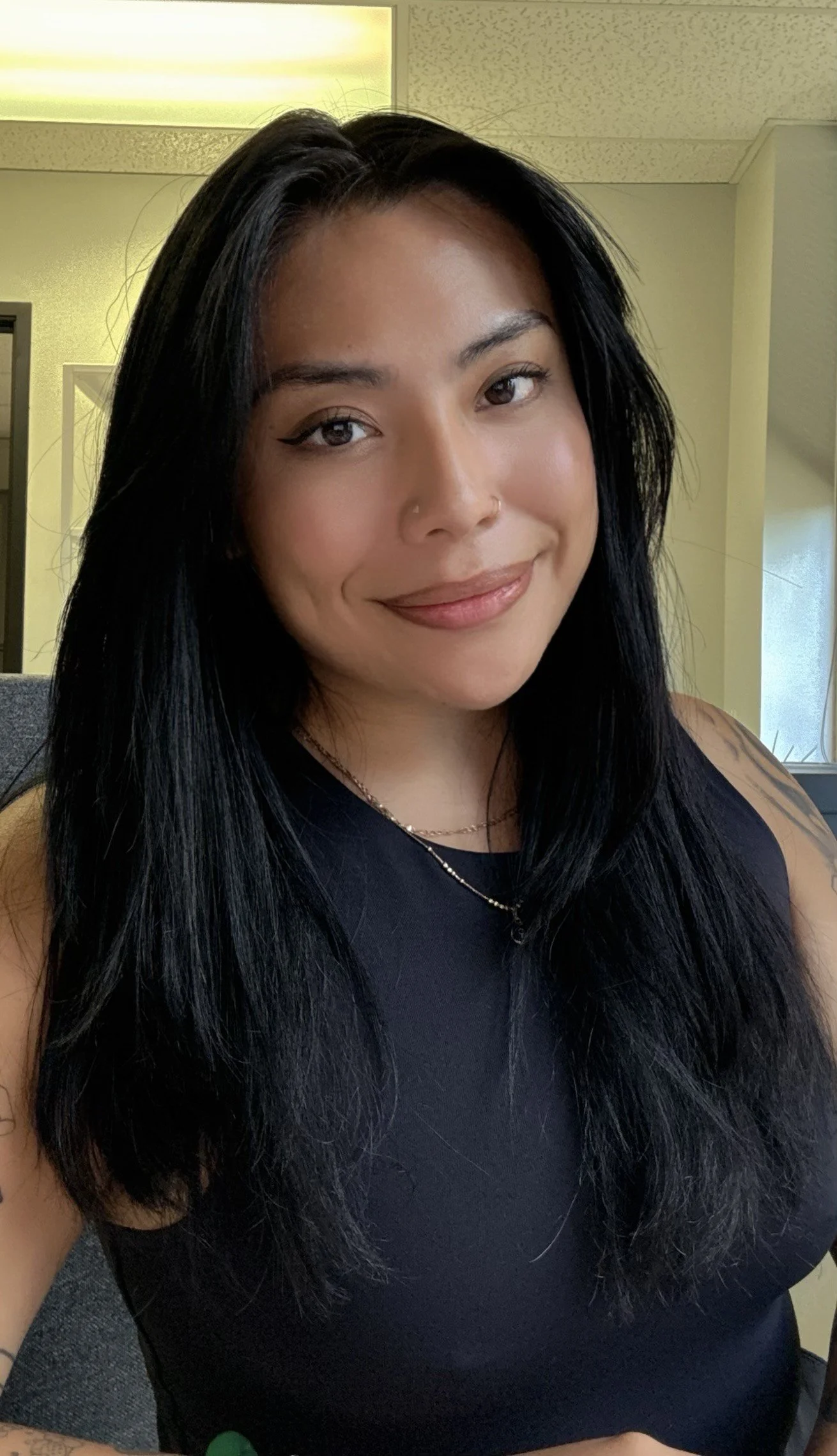 A woman with long black hair, wearing a black top and gold necklaces, smiling in an indoor setting with yellow and green walls.