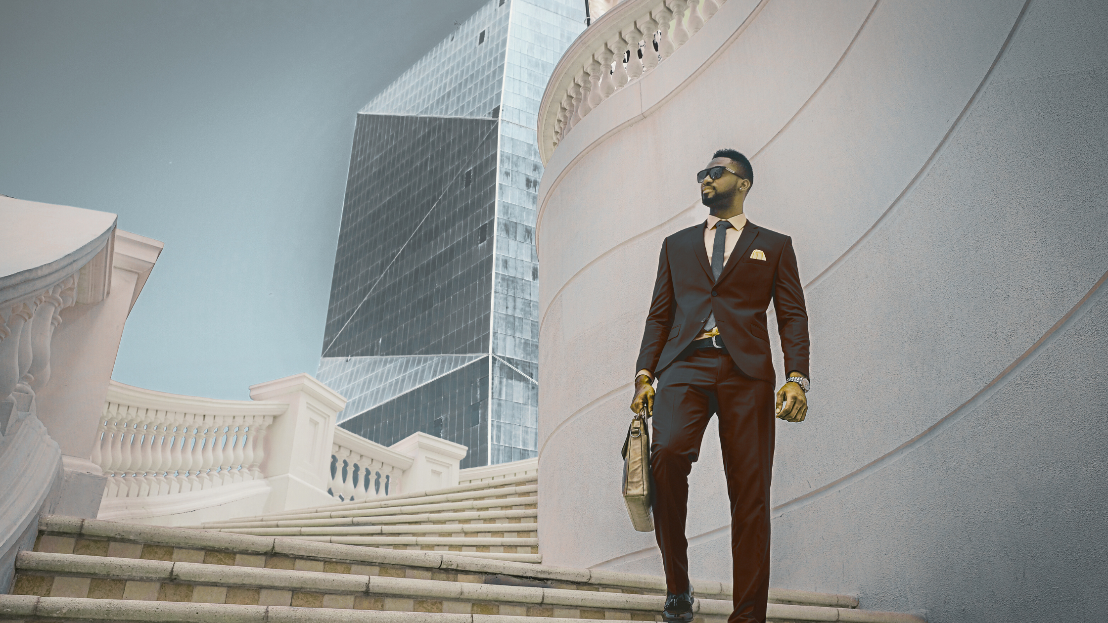 A man in a black suit, sunglasses, and a tie walking down a staircase outside in an urban area, with modern glass buildings in the background.
