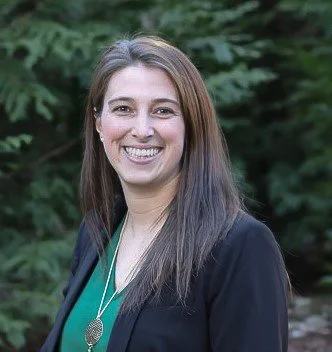 Smiling woman in outdoor setting with trees, wearing a green top and black blazer.