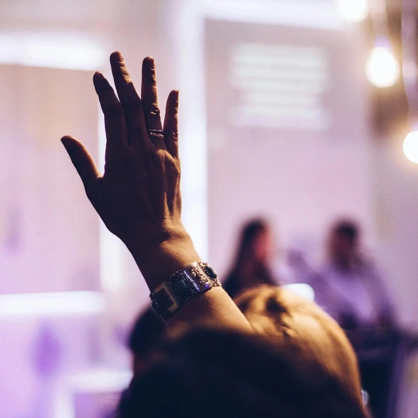 A person raising their left hand during a meeting or event, wearing a watch and rings, with a blurred background of people in a room with purple and white lighting.