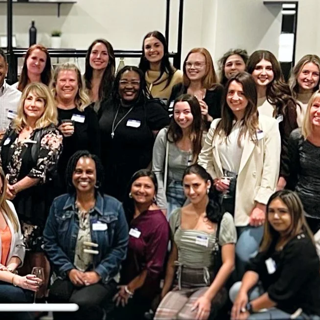 Group of diverse women at a social event, smiling and standing together in an indoor setting.