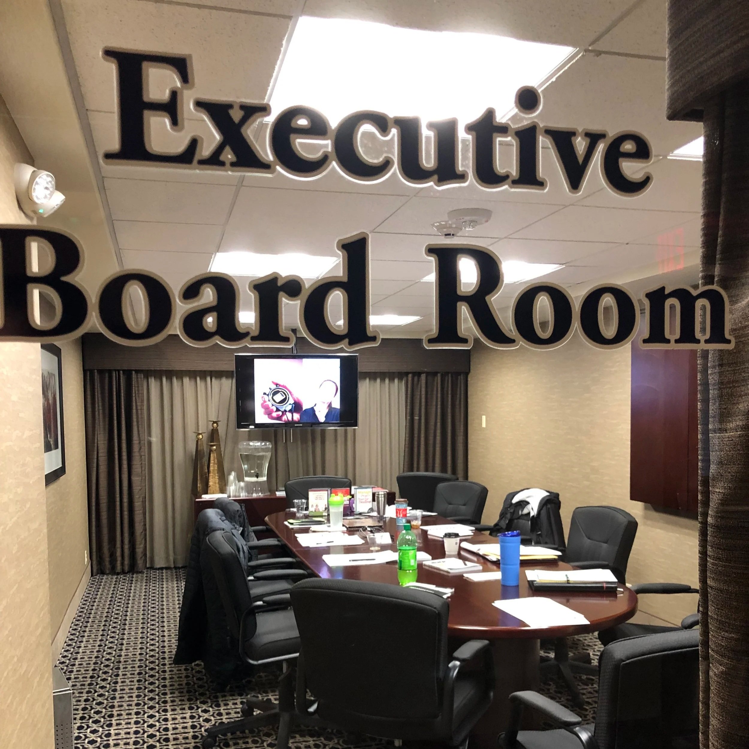 A conference room with a large oval table, chairs, a TV monitor, and bottled drinks. The room is seen through a glass door with a sign that reads 'Executive Board Room'.