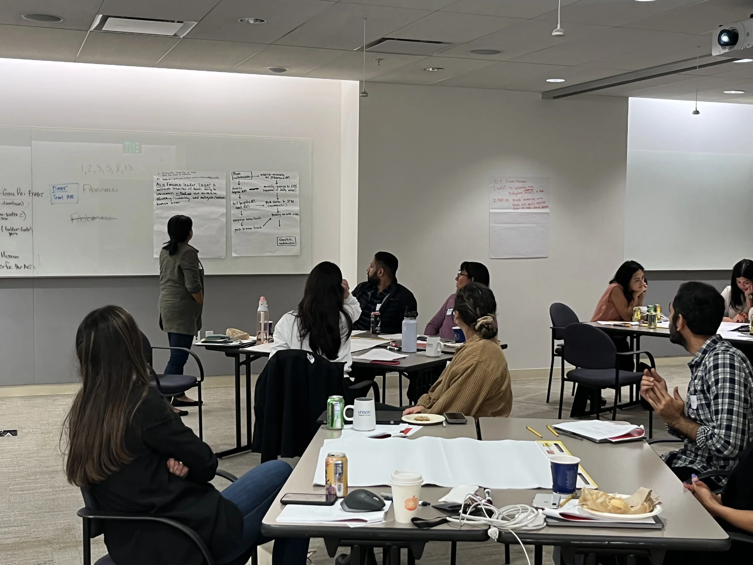 A group of people attending a presentation in a conference room. A woman is standing and speaking near whiteboards with handwritten notes, while seated participants listen and take notes at tables with laptops, notebooks, drinks, and snacks.