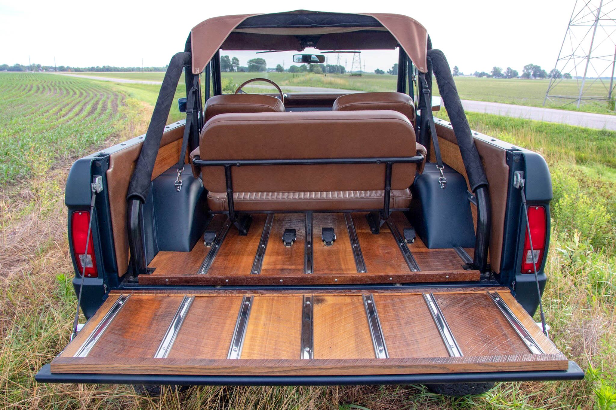 Open tailgate of a vintage SUV with wood paneling and exposed seating, parked on a grassy field road.
