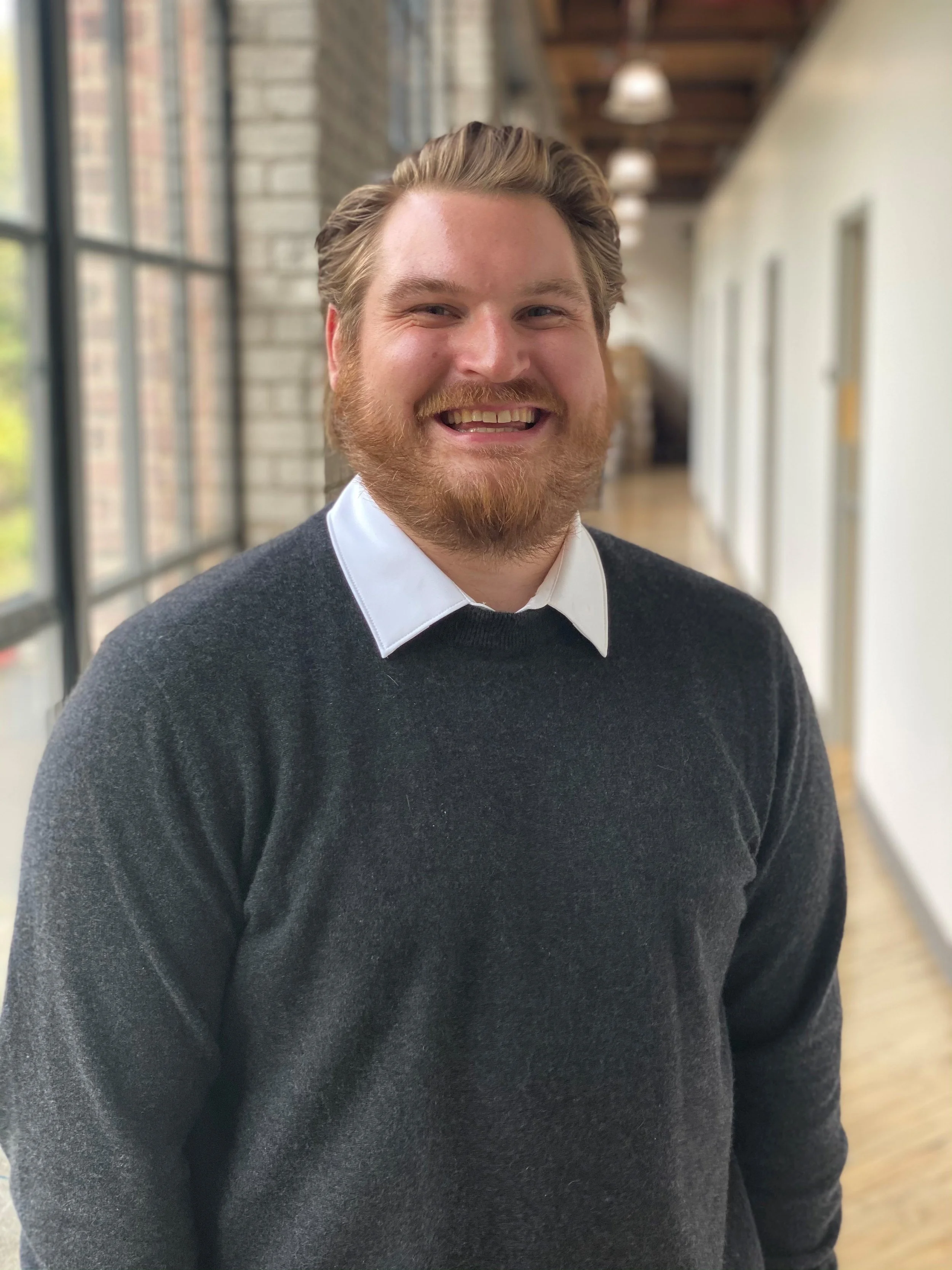 A smiling man with a beard and mustache, wearing a white collared shirt and a dark gray sweater, standing in a bright indoor hallway with large windows and brick walls.