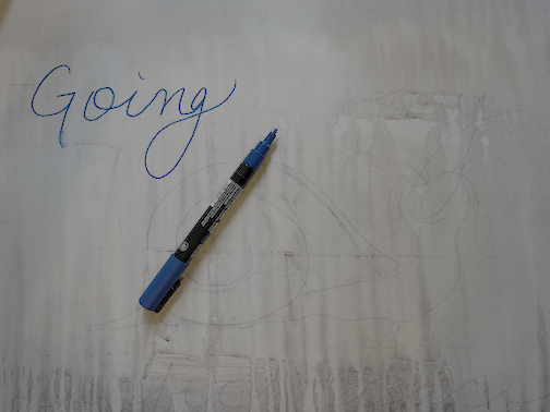 The word 'Going' written in blue ink on paper with a blue and black pen resting on the paper.