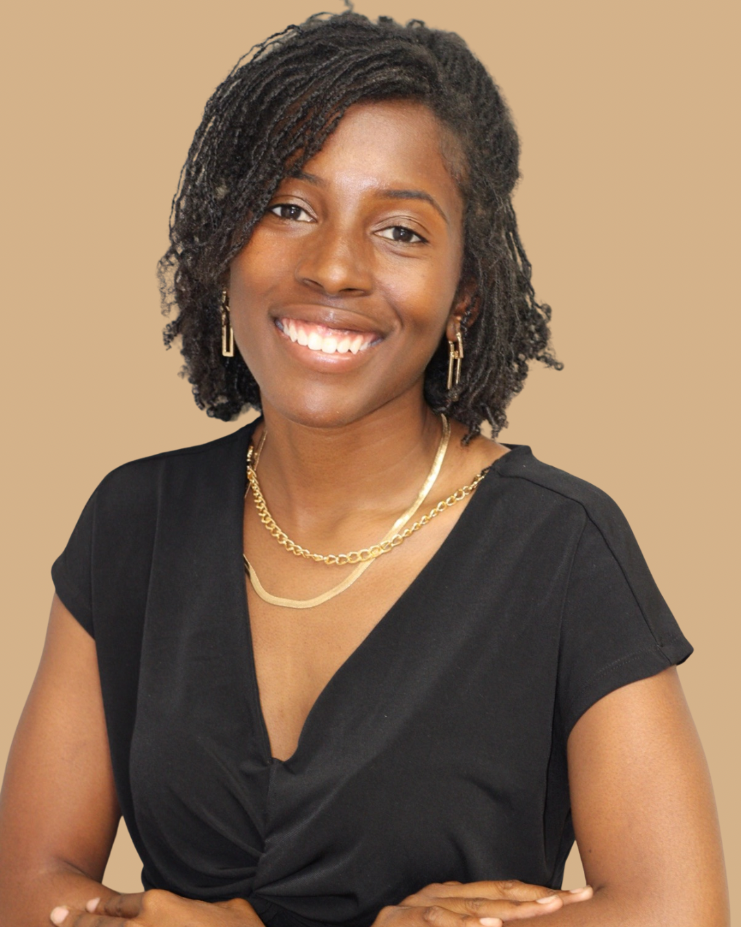 A smiling woman with dark, curly, shoulder-length hair wearing a black top and gold jewelry, posing against a beige background.
