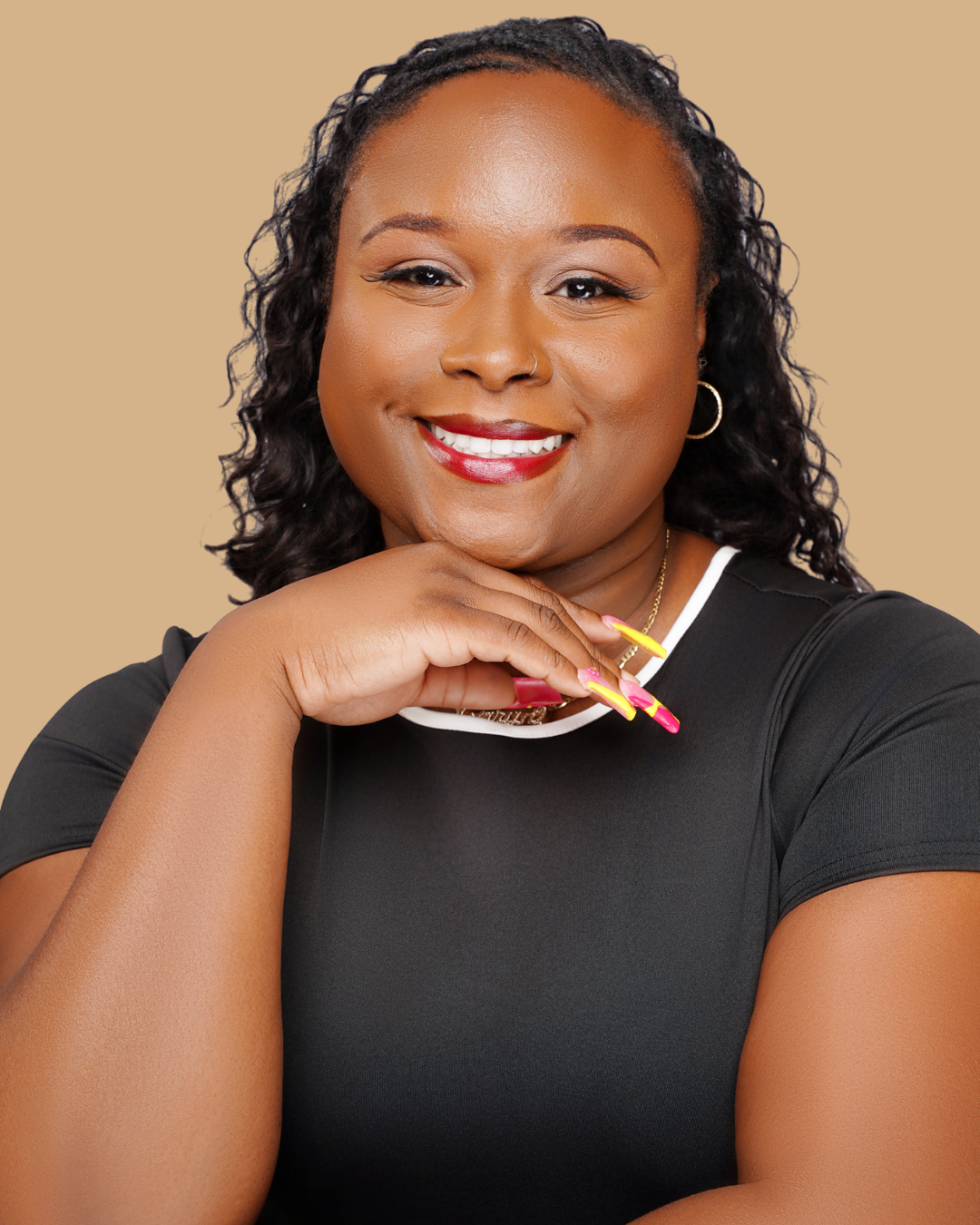 A woman with dark curly hair smiling, wearing a black top with white trim, gold hoop earrings, and colorful pink and yellow nails against a beige background.