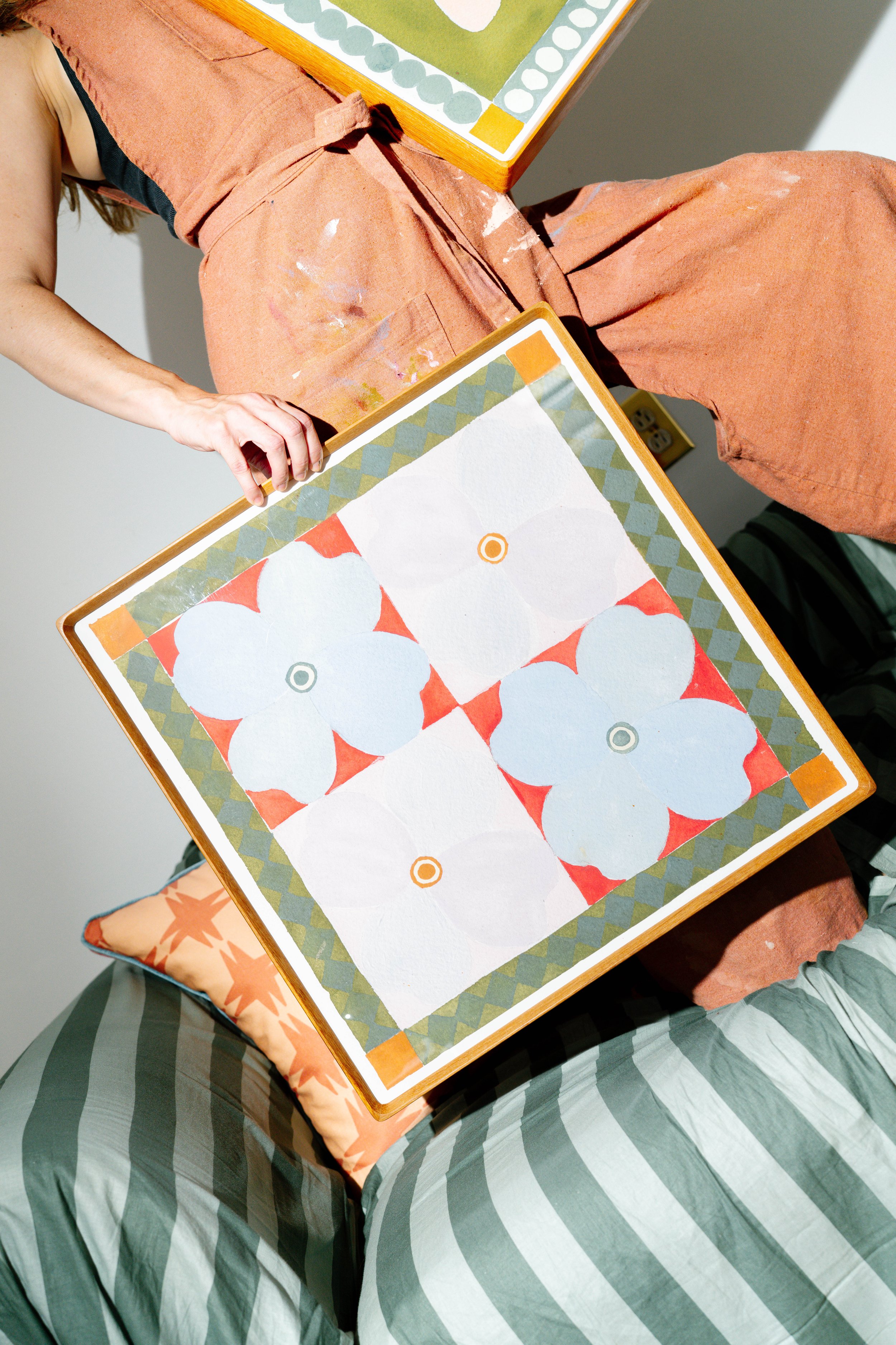 Person sitting on a cushioned surface, holding a colorful geometric and floral patterned artwork. The person wears apricot-colored work clothes with paint splatters and striped pants. The artwork features four large white flowers with orange centers,
