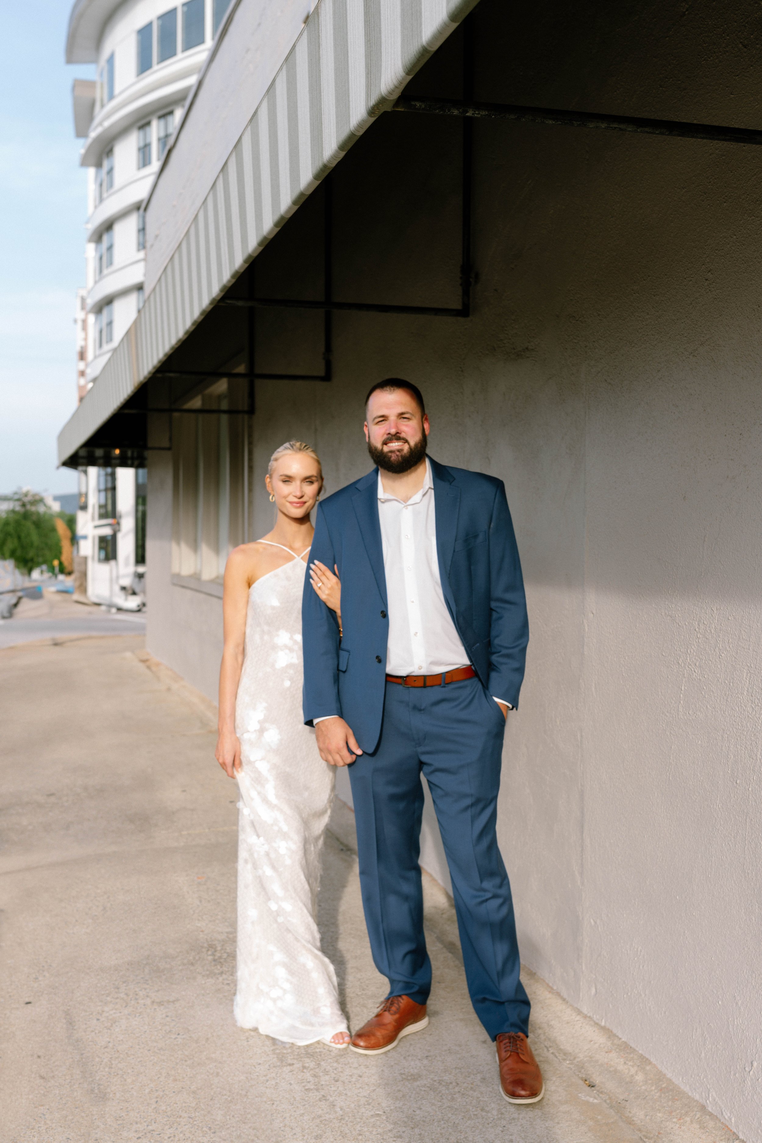 A couple posing outdoors; a woman in a white dress and a man in a blue suit stand close together.