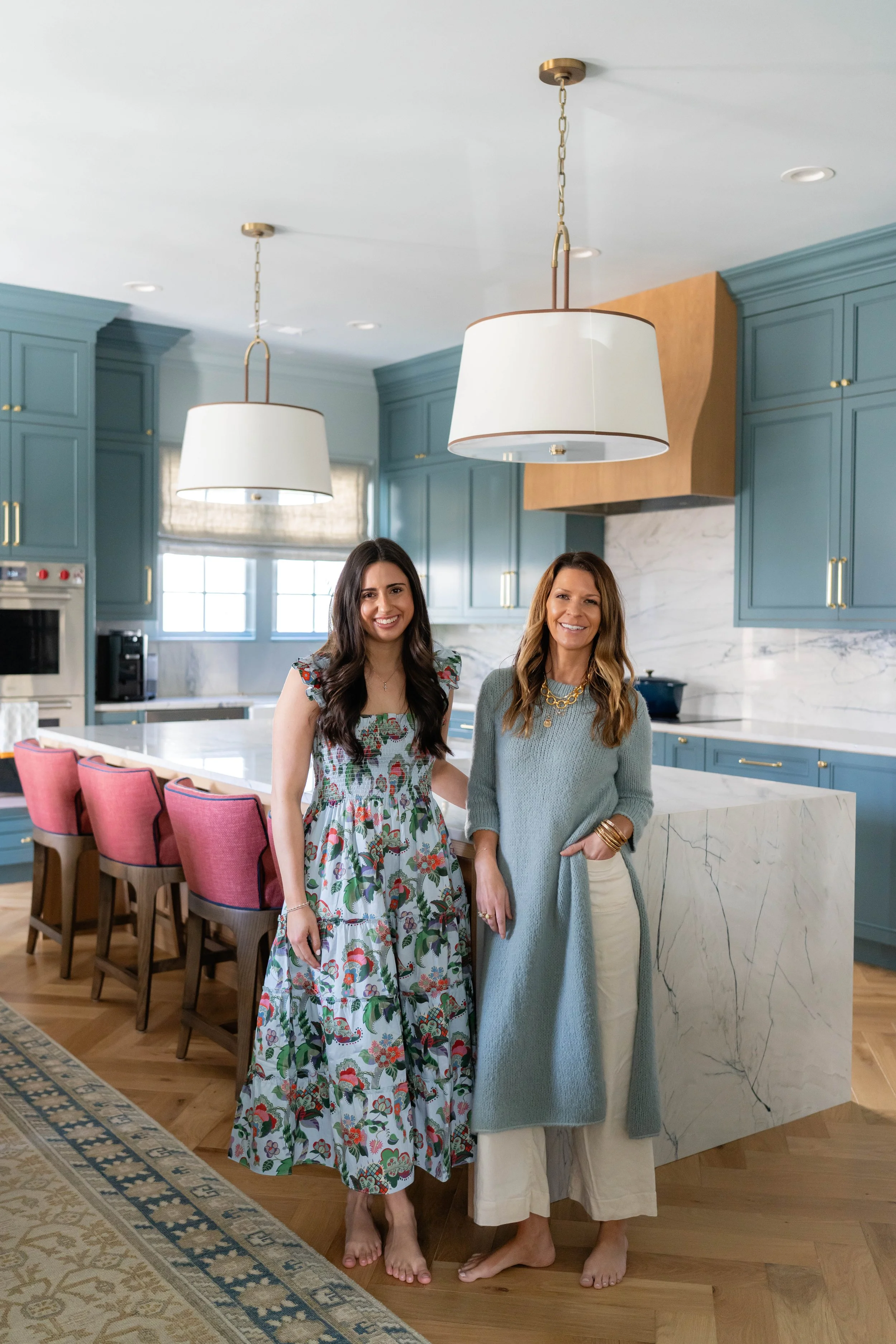 Two women smiling and standing barefoot in a modern kitchen with blue cabinets, a marble island, and pendant lights.