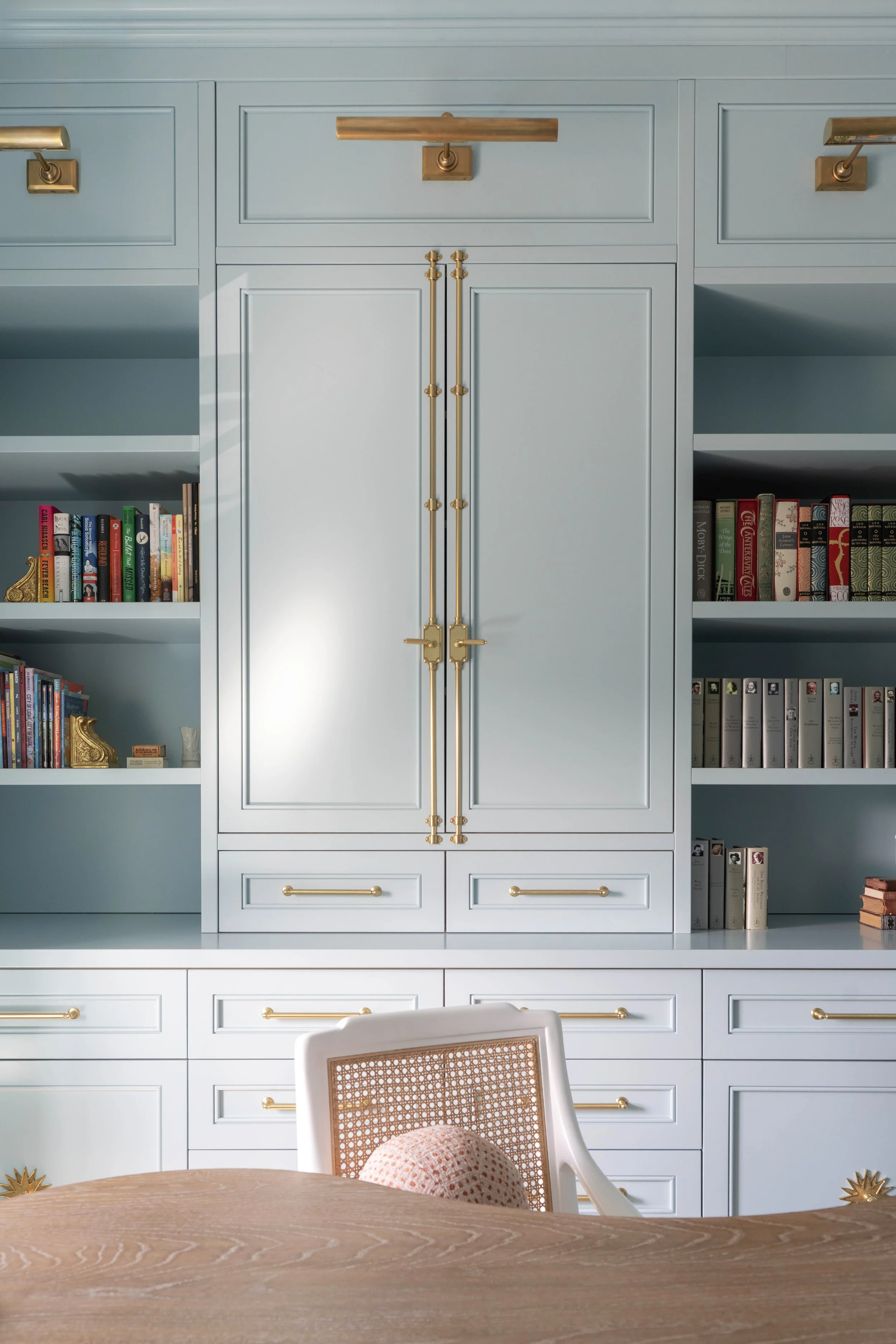 A built-in light blue cabinet with gold handles and accents, housing books and decorative items, visible from the back of a dining room with a wooden table and a white chair with a woven backrest.