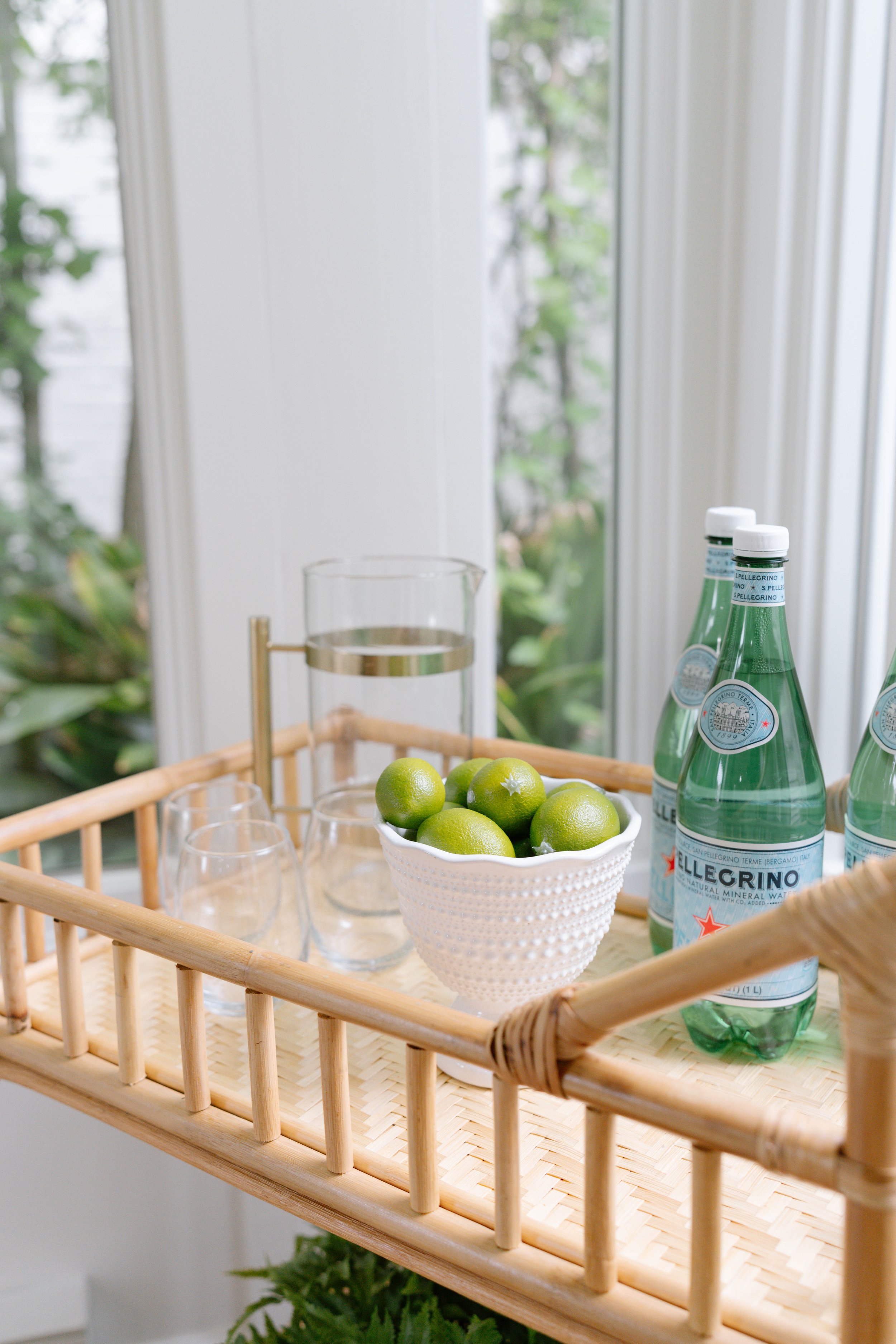 Bamboo cart with San Pellegrino bottles, white bowl of limes, glass pitcher, and empty glasses in a bright room.