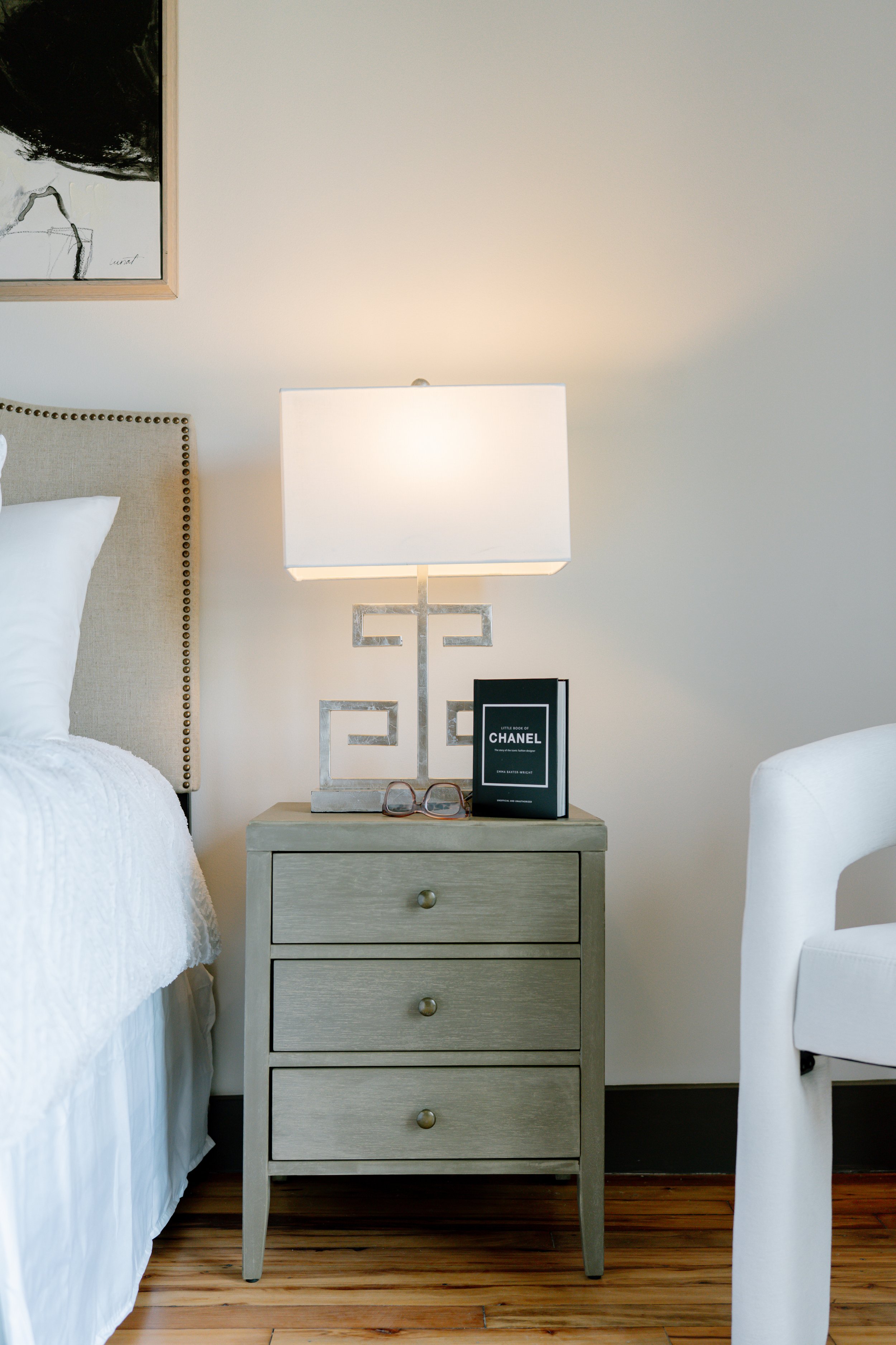 Styled bedroom vignette with a gray three-drawer nightstand, Greek key table lamp, and fashion book beside a linen nailhead headboard and white accent chair, captured during a commercial interior photography session in downtown Columbia, SC