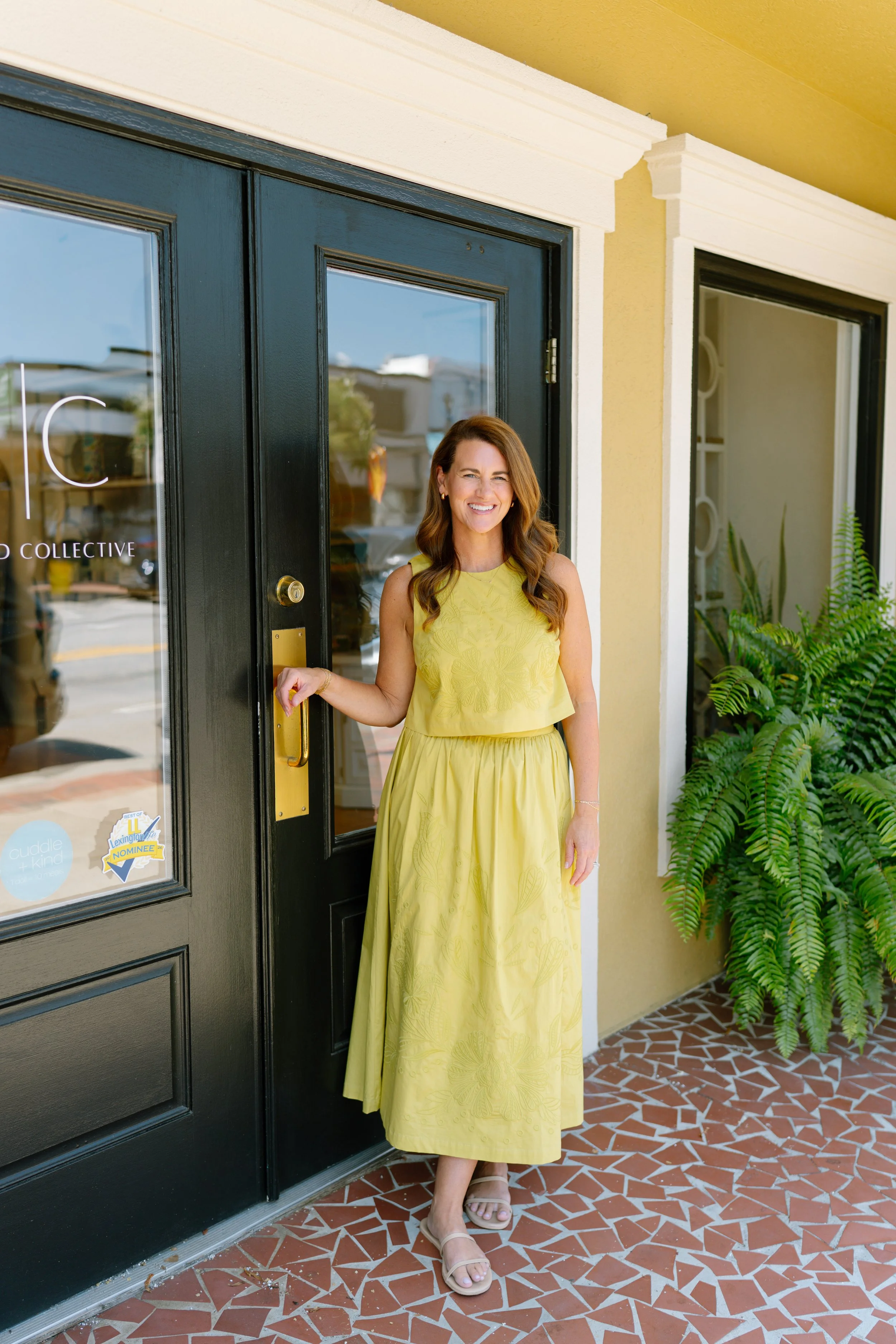 Smiling owner of Tenfold Collective in a yellow embroidered two-piece set standing at the boutique’s black front doors, with a fern and mosaic tile entry, captured during a commercial brand photography session in Lexington, SC