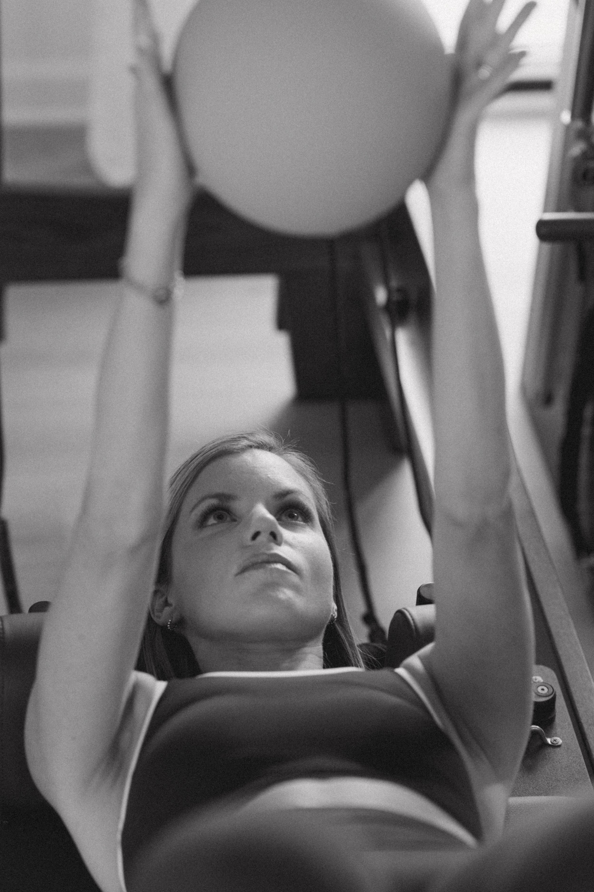 Black and white image of a woman lying on a Pilates reformer holding a small exercise ball overhead at Tone Wellness, captured during a commercial fitness photography session for a Pilates studio in Lexington, SC