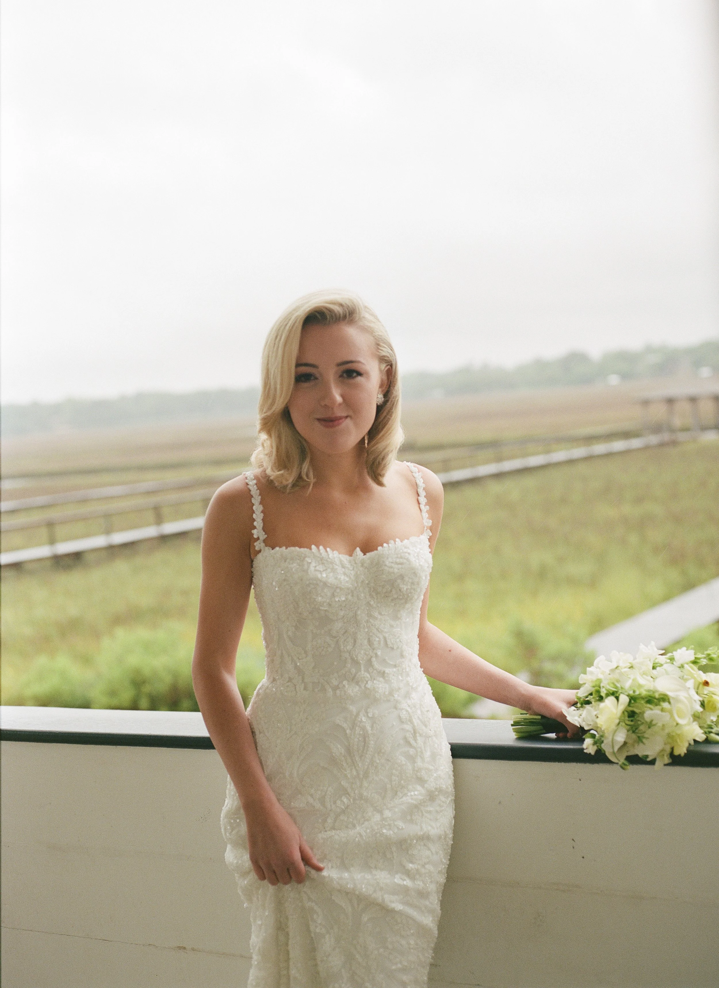 Bride in lace fitted wedding gown holding white bouquet on marsh-view balcony, Charleston SC wedding photographer