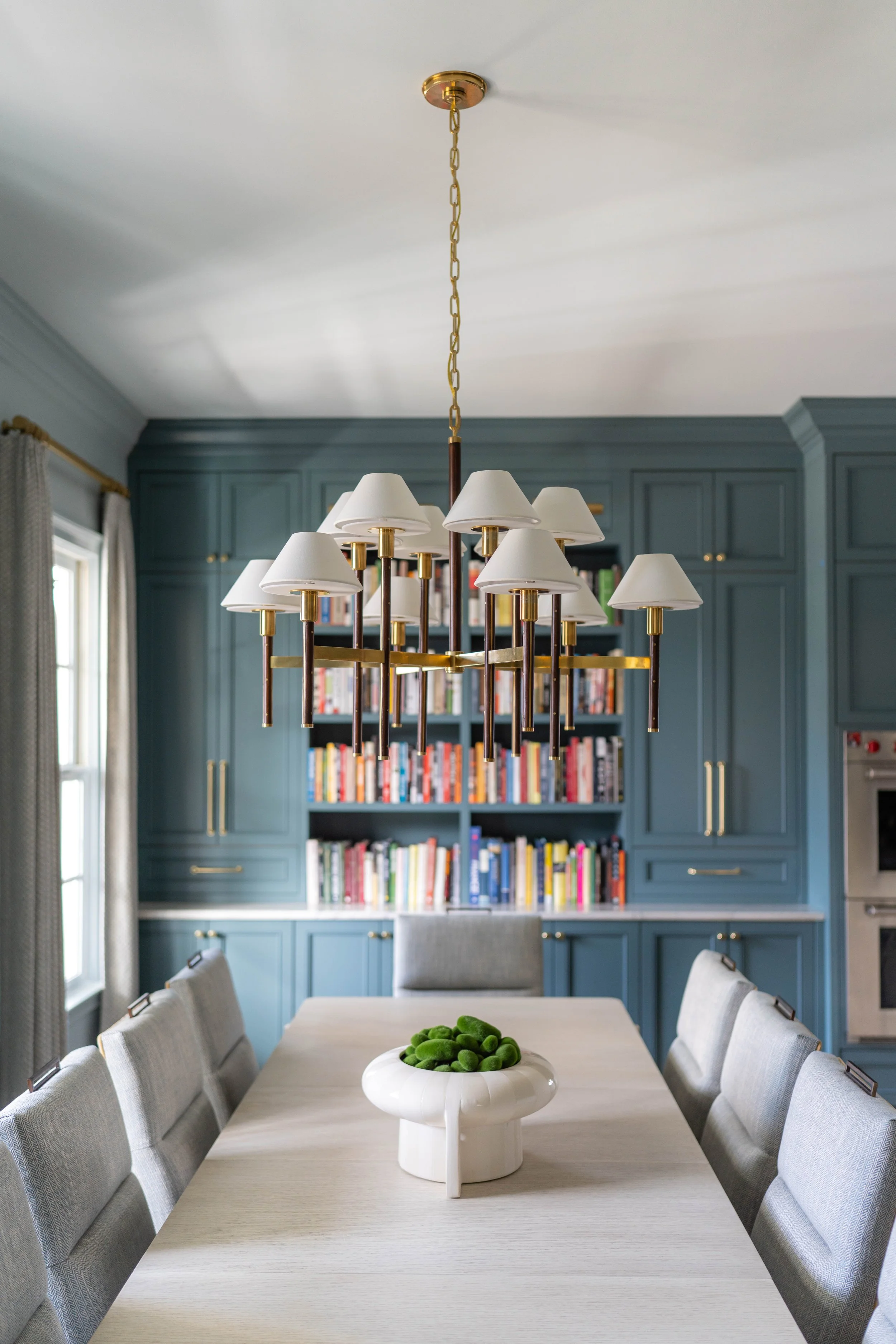 Modern dining room with a light-colored table, upholstered chairs, a white vase with green decorative objects, and a navy blue built-in bookshelf filled with books, illuminated by an elegant chandelier.