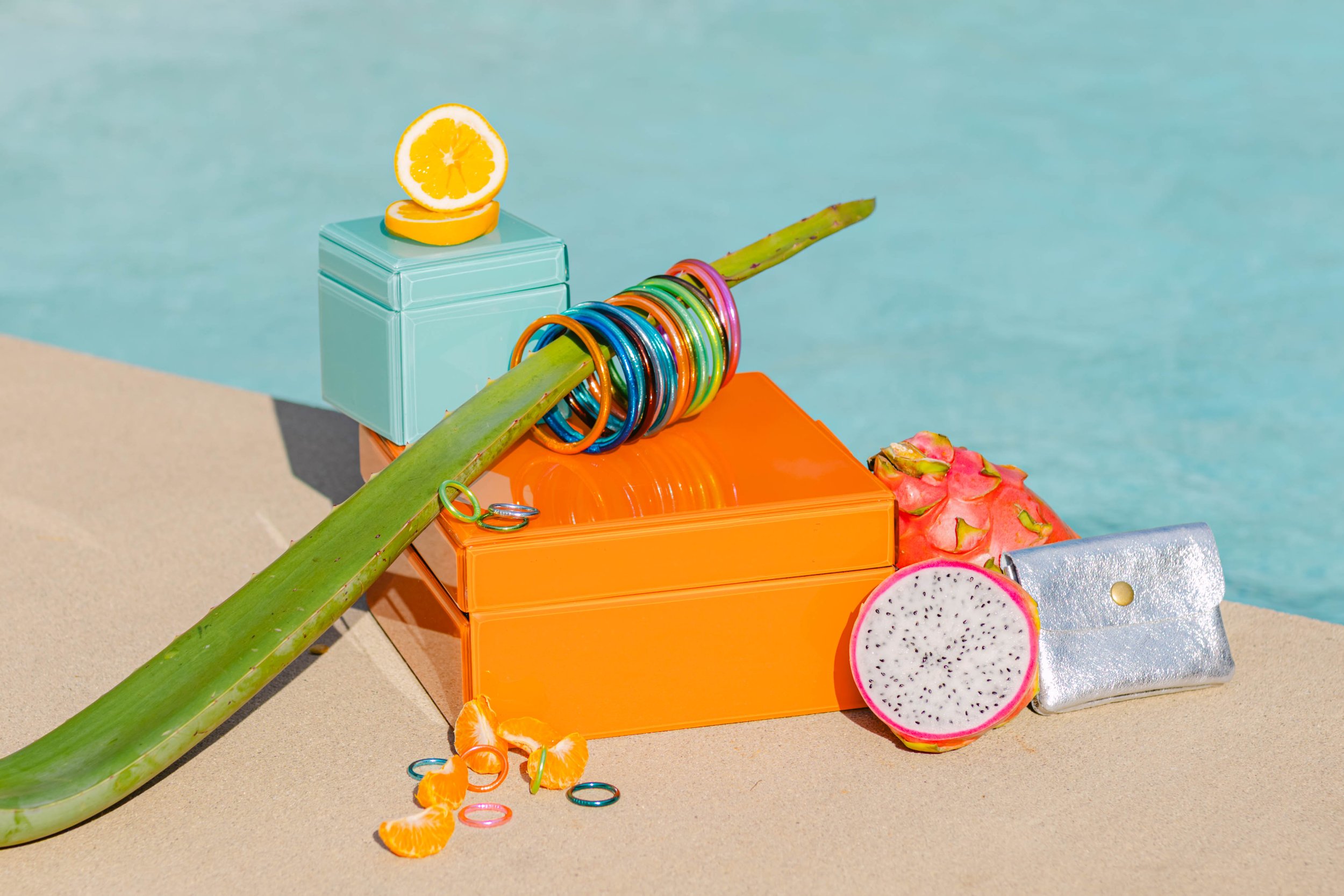 Colorful jewelry and accessories arranged on a beach-side surface with a swimming pool in the background, including bracelets, rings, a small purse, and sliced tropical fruits.